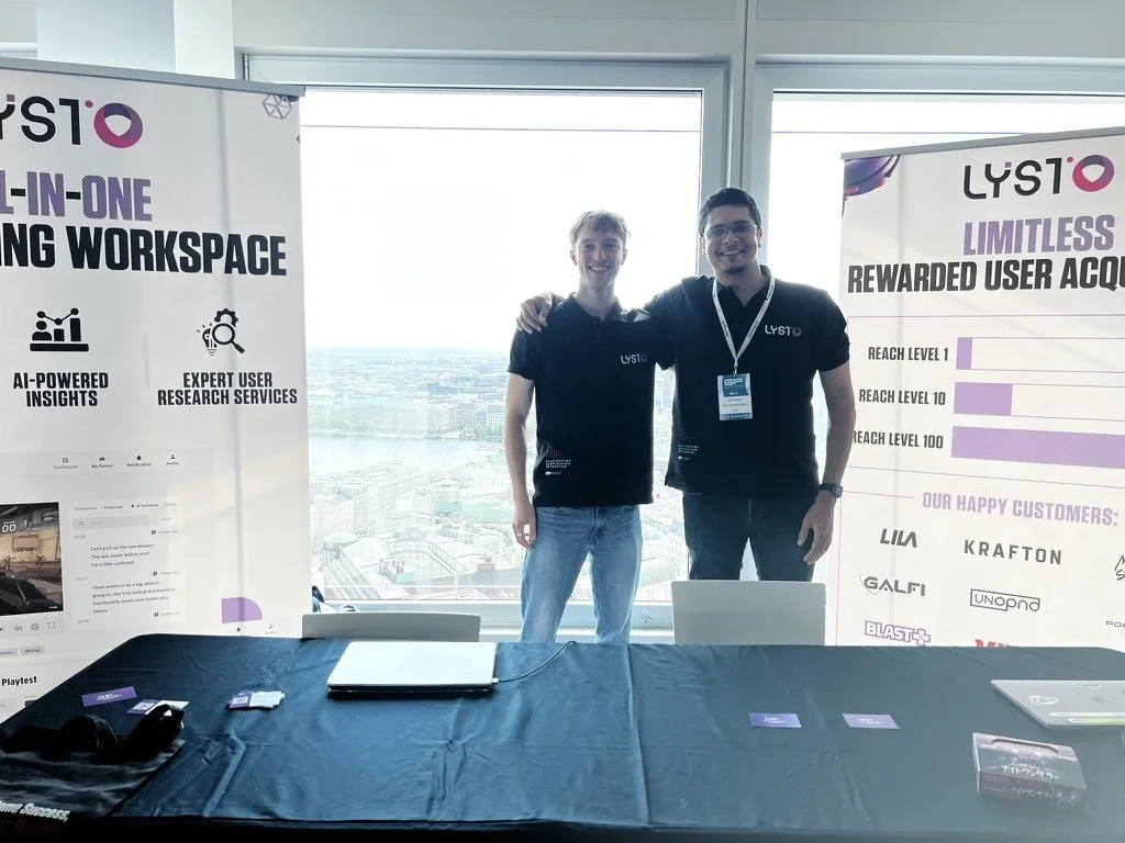 Two men standing behind a table at a conference booth, both wearing black shirts with the 'LYSTO' logo, posing with their arms around each other's shoulders. The booth features banners promoting LYSTO's all-in-one workspace solutions, with the city skyline visible through large windows in the background.