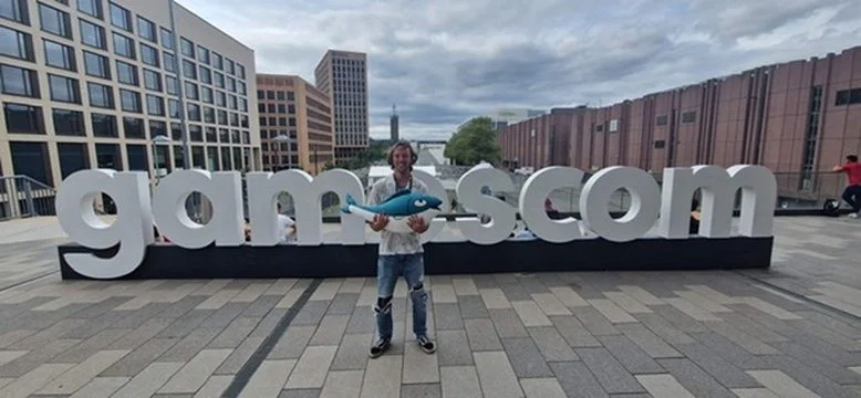 A man holding a large blue fish prop in front of large white letters spelling 'gam-scom' on a rooftop with city buildings in the background.