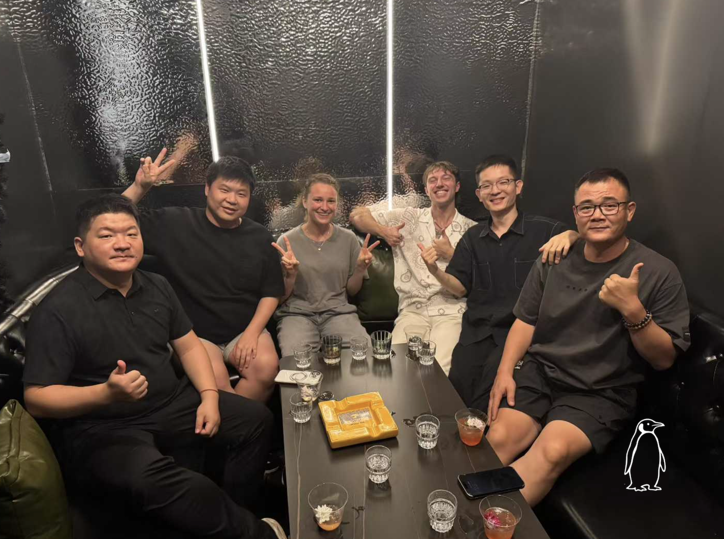 Group of six people sitting around a black table in a dark room, smiling and making peace signs and thumbs-up gestures. There are glasses and small plates on the table, and a yellow ashtray.