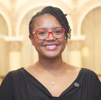 Dr. Watt - A woman with glasses and a black top smiling in front of a patterned background.