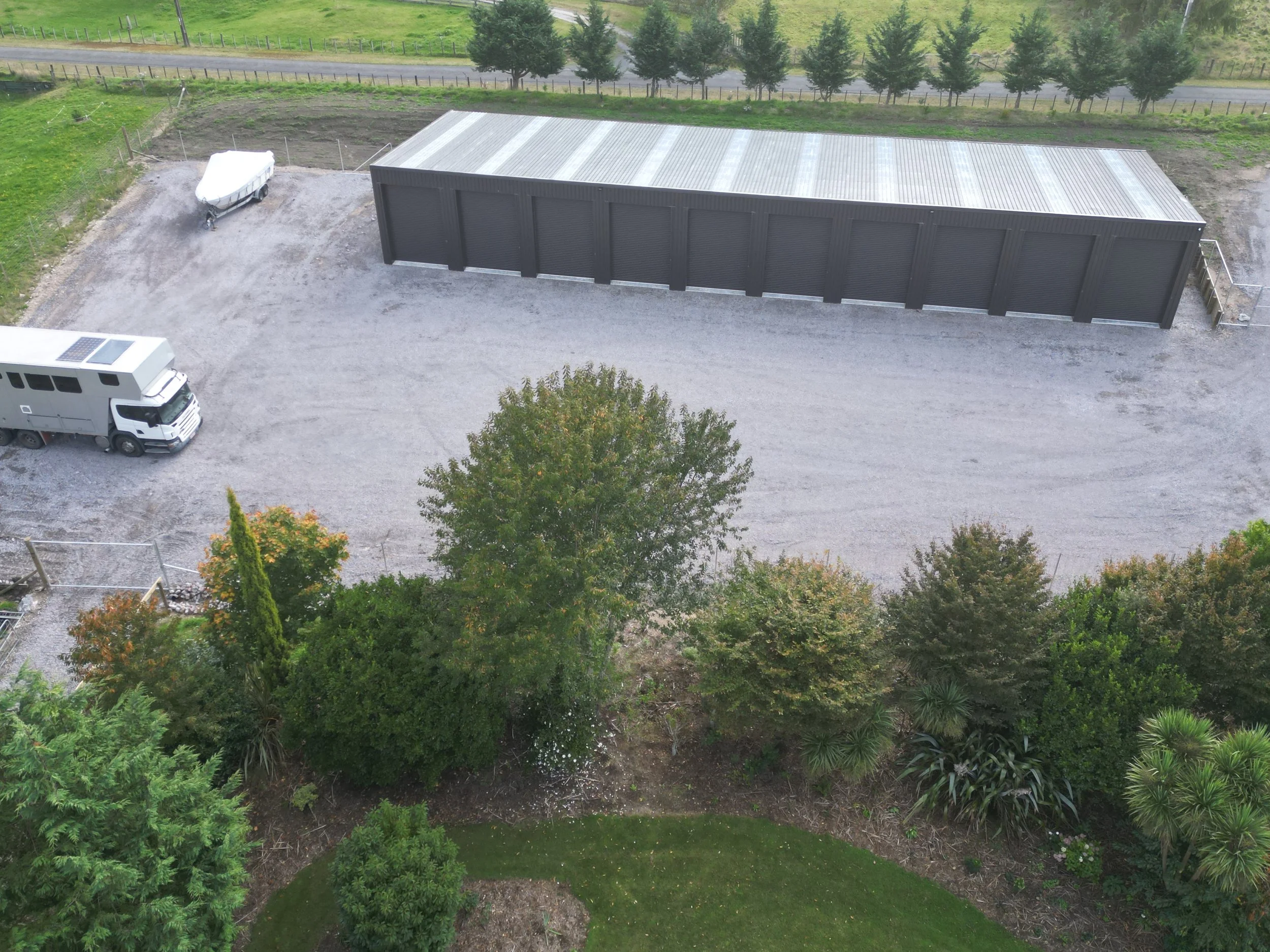 An aerial view of a gravel parking area with a white RV, a small boat, a large black storage shed, and some trees and greenery nearby.