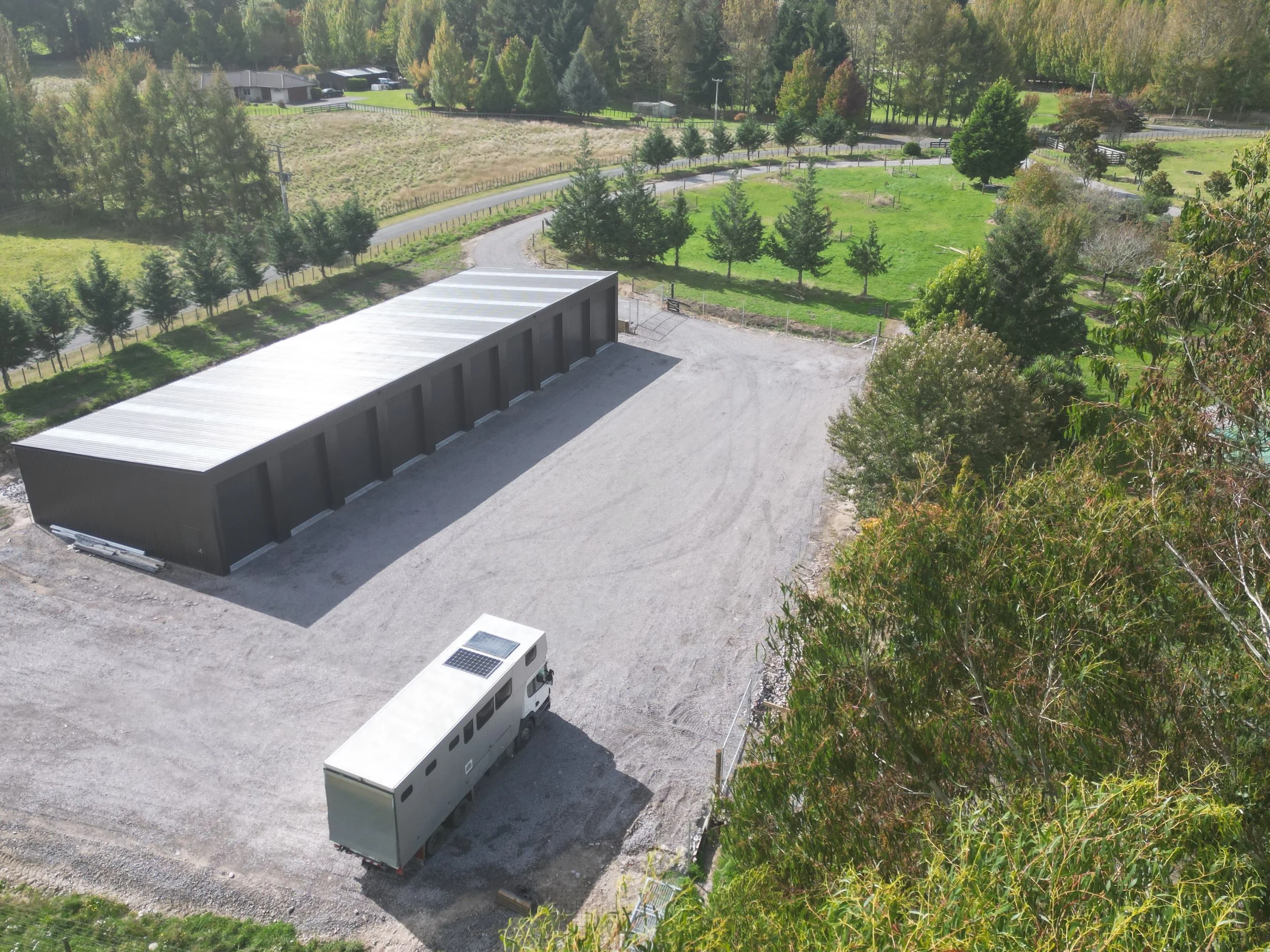 An aerial view of a rural property showing a large metal storage building, a small white trailer with solar panels, and a gravel driveway surrounded by green trees and grassy fields.