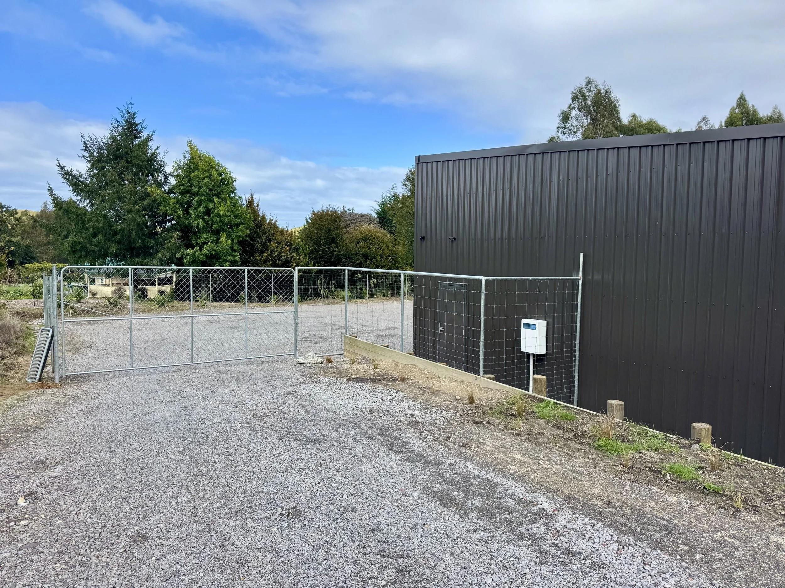 Exterior view of a gravel driveway with a chain-link gate, black metal building, and greenery in the background under a partly cloudy sky.