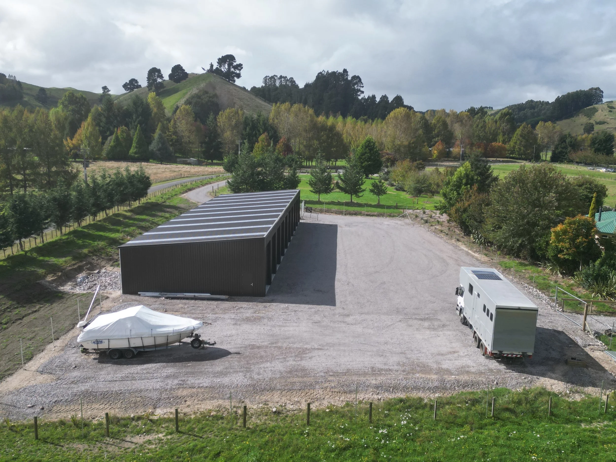 Large black metal building with a curved roof, gravel parking area with a boat covered in white tarp on a trailer, and a white mobile home with a solar panel on the roof, set in a rural landscape with trees and hills in the background.