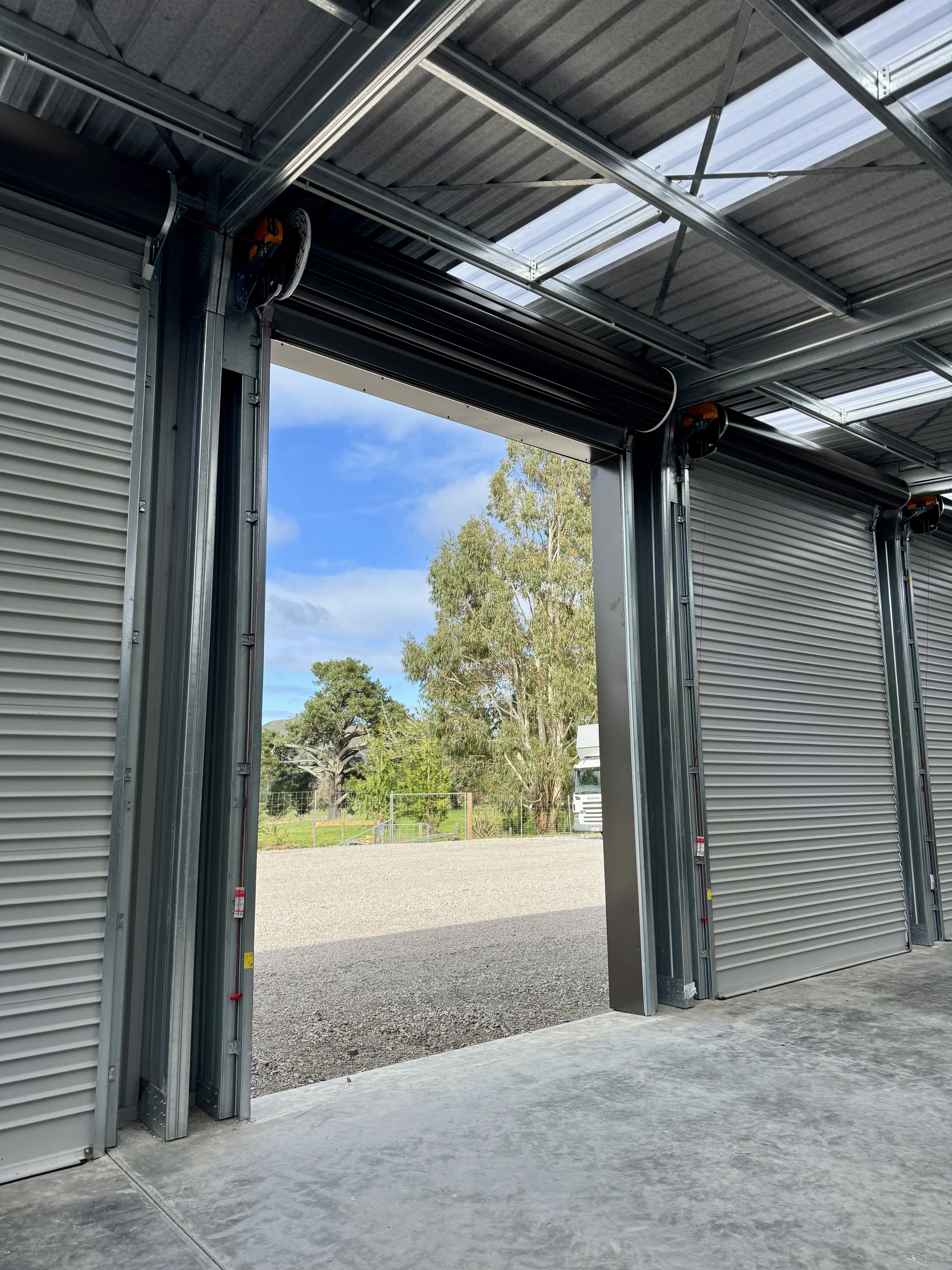 Open roll-up garage doors revealing a gravel yard with green trees and blue sky outside.