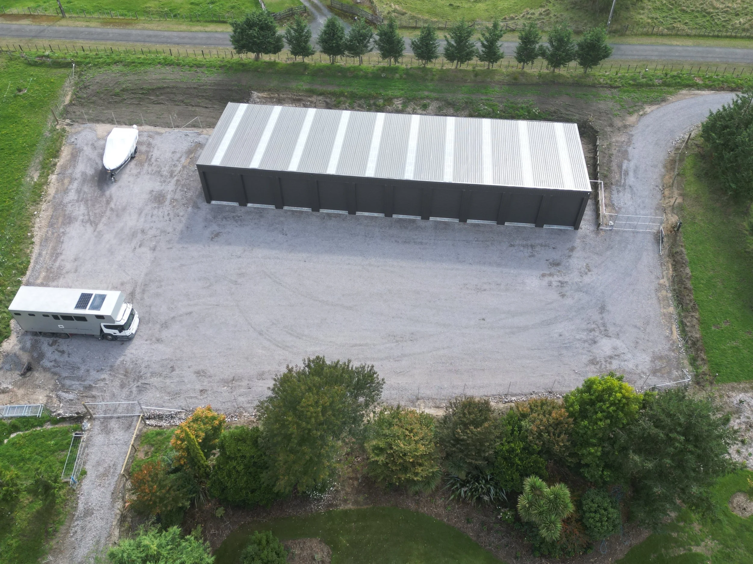 Aerial view of a large fenced gravel lot with a black metal storage container, a white truck with solar panels, and a boat, surrounded by green trees and a lawn.