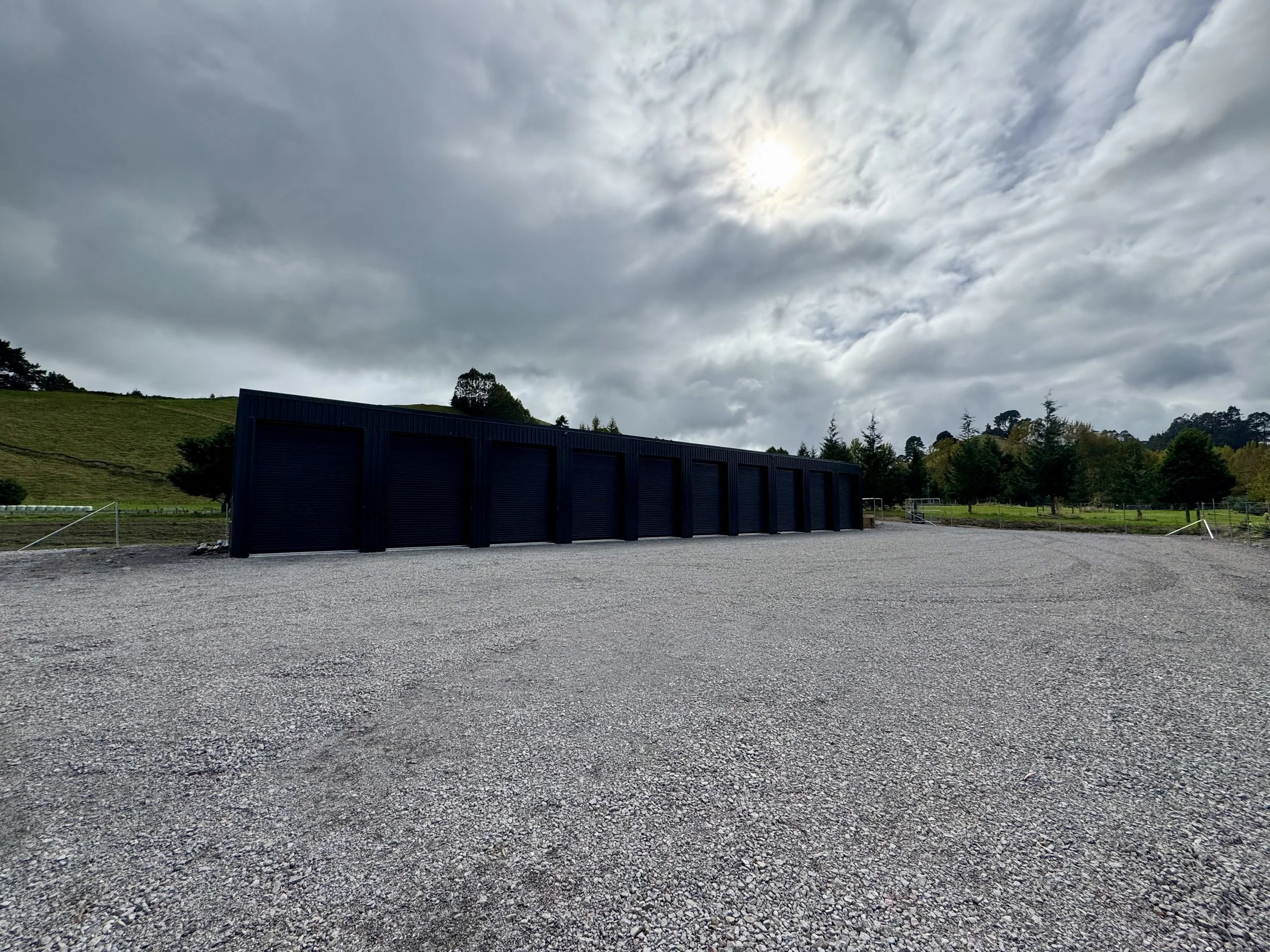 A black storage unit with multiple roll-up doors located on a gravel lot with a cloudy sky overhead and trees in the background.