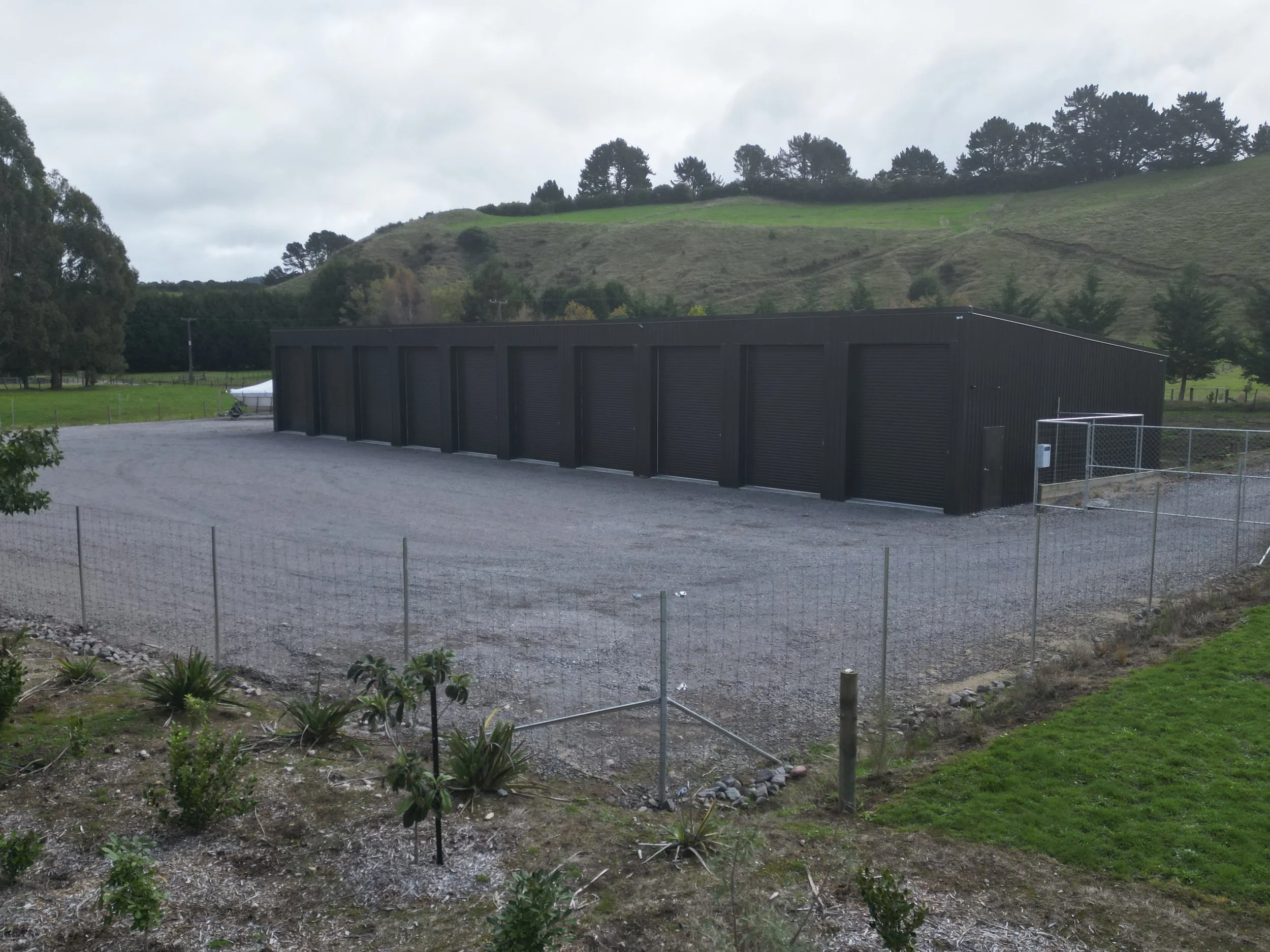 A black storage unit with multiple roll-up doors, set in an open lot with gravel ground, surrounded by a chain-link fence, with a grassy field and rolling hills in the background under a cloudy sky.