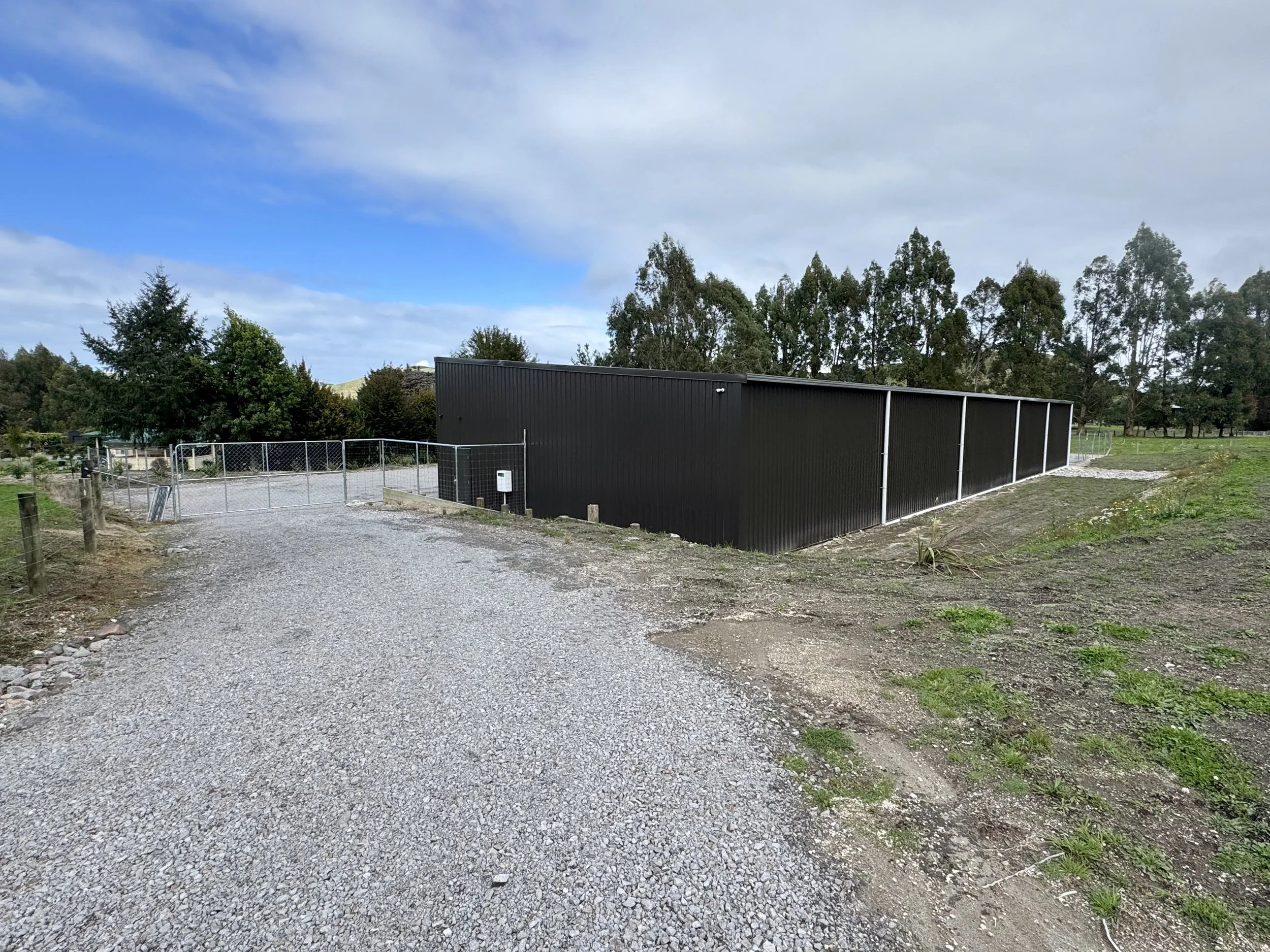 A black metal structure on a gravel path with a chain-link fence and a gate. Surrounding trees and vegetation under a partly cloudy sky.