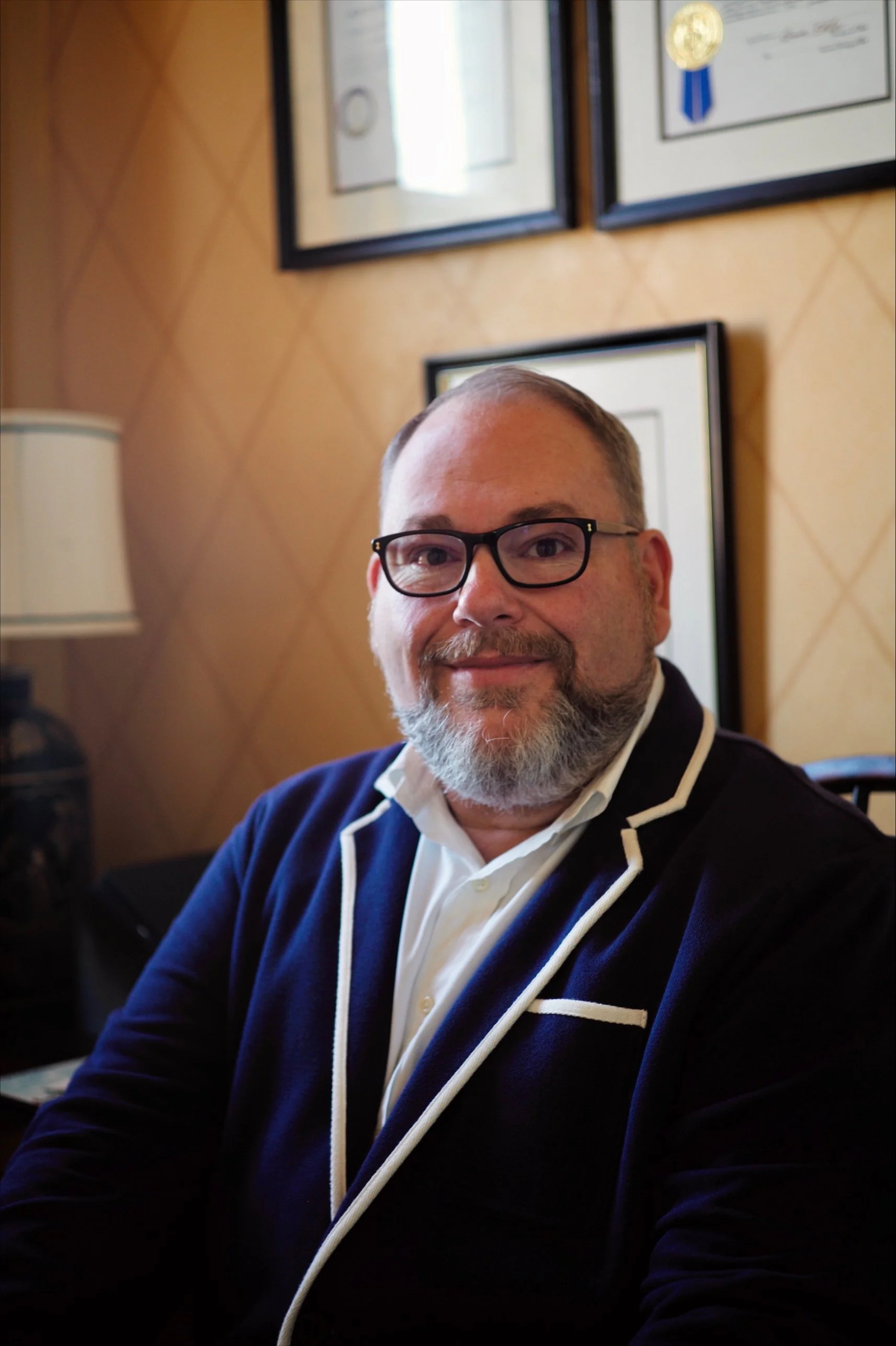 A man with glasses and a beard wearing a navy blazer with white piping and a white shirt, sitting indoors with framed certificates on the wall behind him. Nathan Saucedo, Kentucky, Kentucky House of Representatives, 55th District