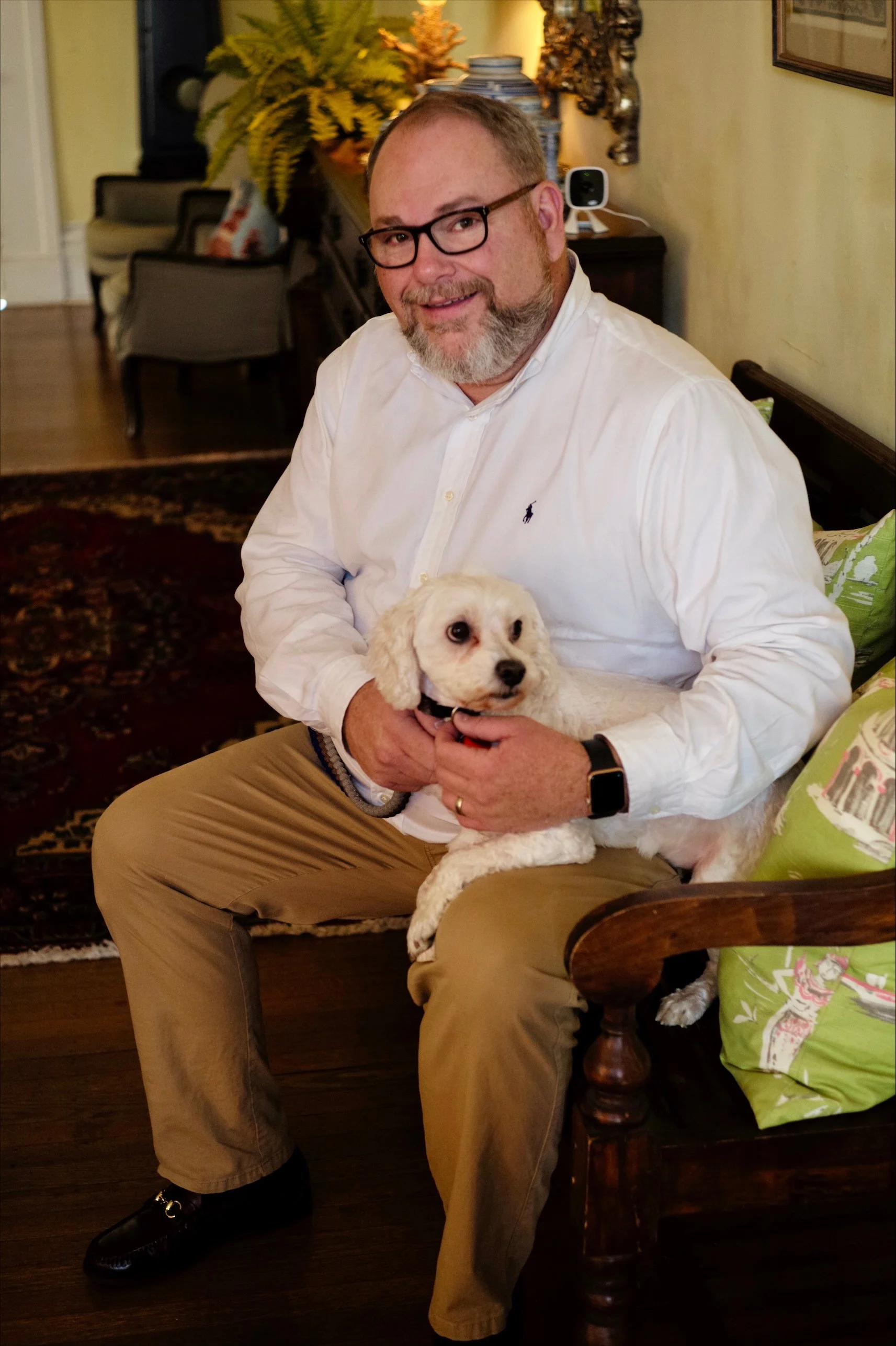 A man with glasses, a beard, and a white shirt sitting on a wooden chair, holding a small white dog in his lap, in a cozy, decorated living room. Nathan Saucedo, Kentucky, Kentucky House of Representatives, 55th District