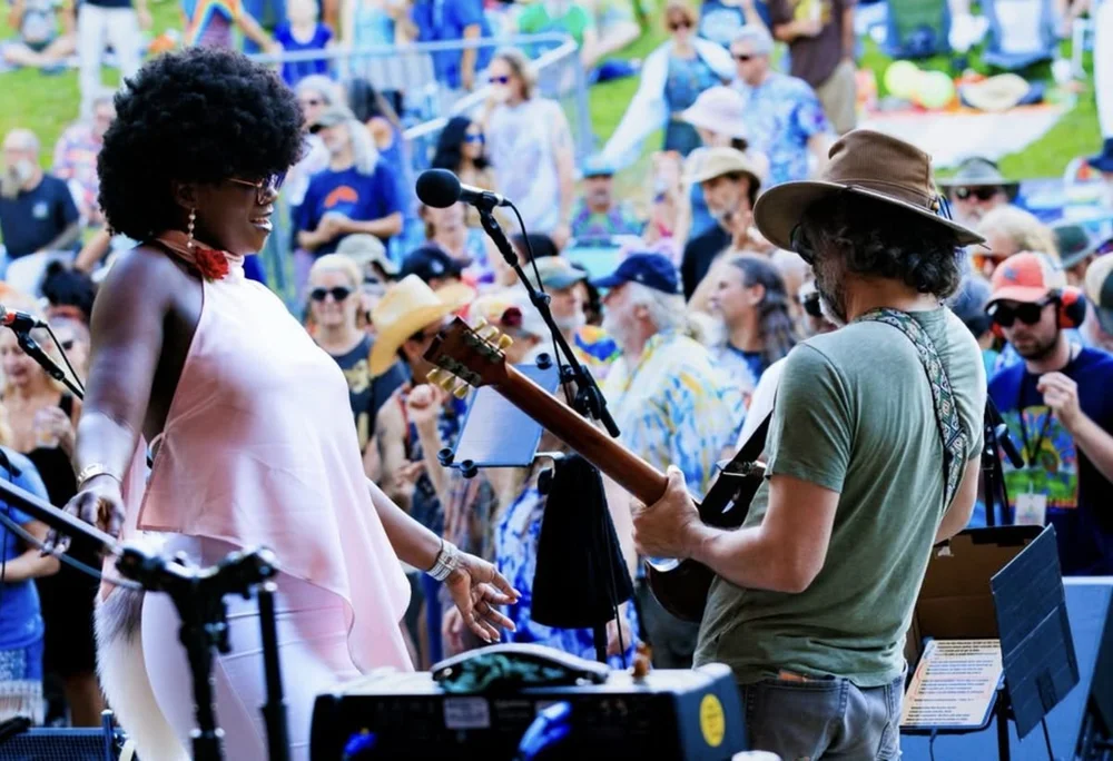 A singer dancing beside a guitarist in a wide-brim hat, crowd behind them