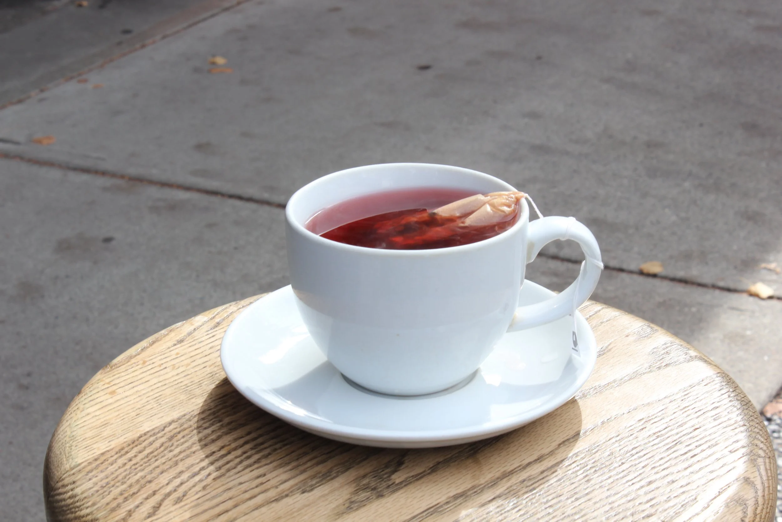 A white ceramic cup filled with hot tea on a matching saucer, placed on a wooden surface outdoors on a sidewalk.