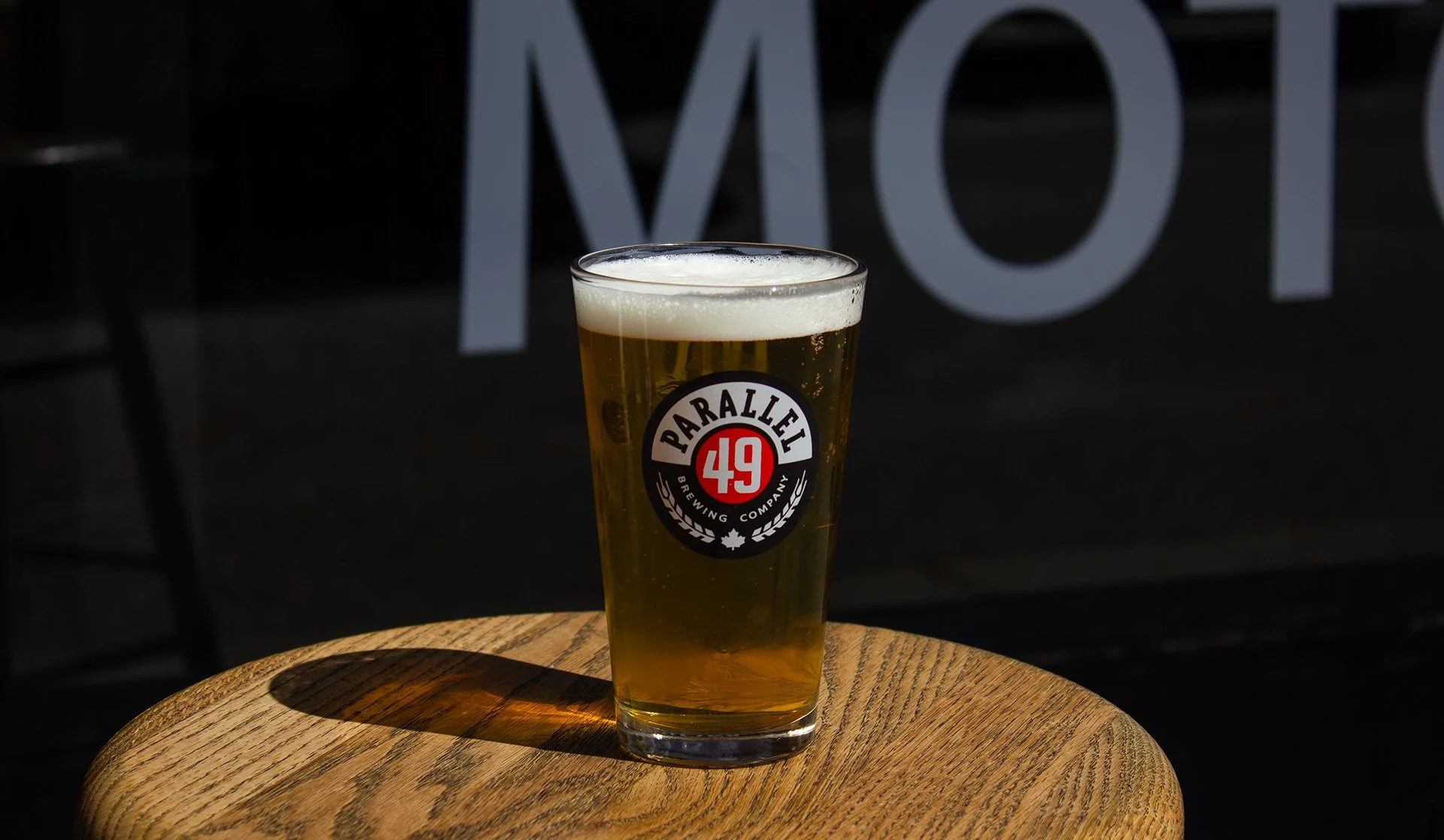 A glass of beer with a frothy head on a wooden table, with a partially visible sign or window in the background.