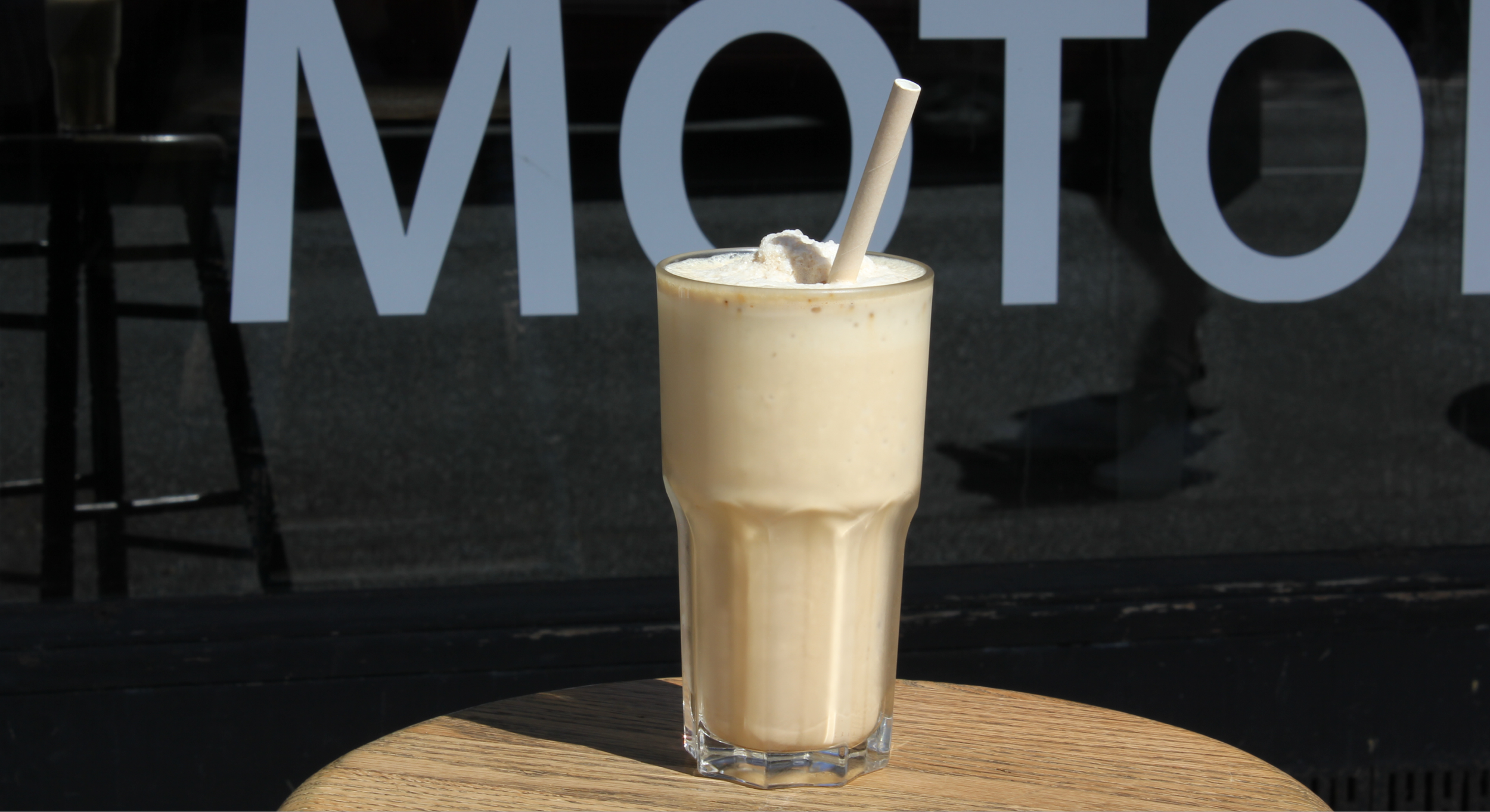 A tall glass of banana smoothie topped with ice cream, with a straw, placed on a wooden table outside a cafe with the word 'MOTO' in the background.