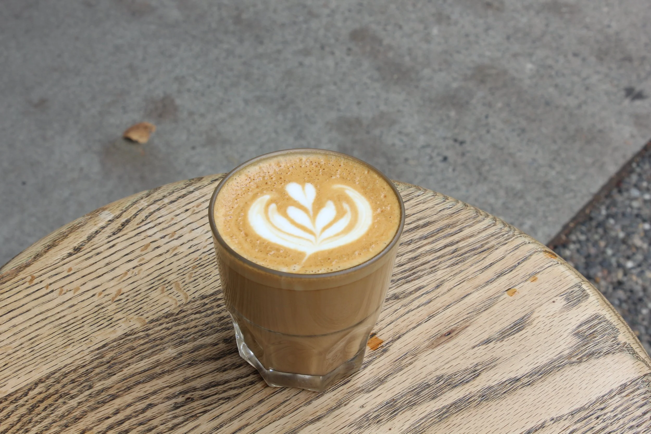 A glass of latte with a heart-shaped foam art on top, placed on a round wooden table outdoors.