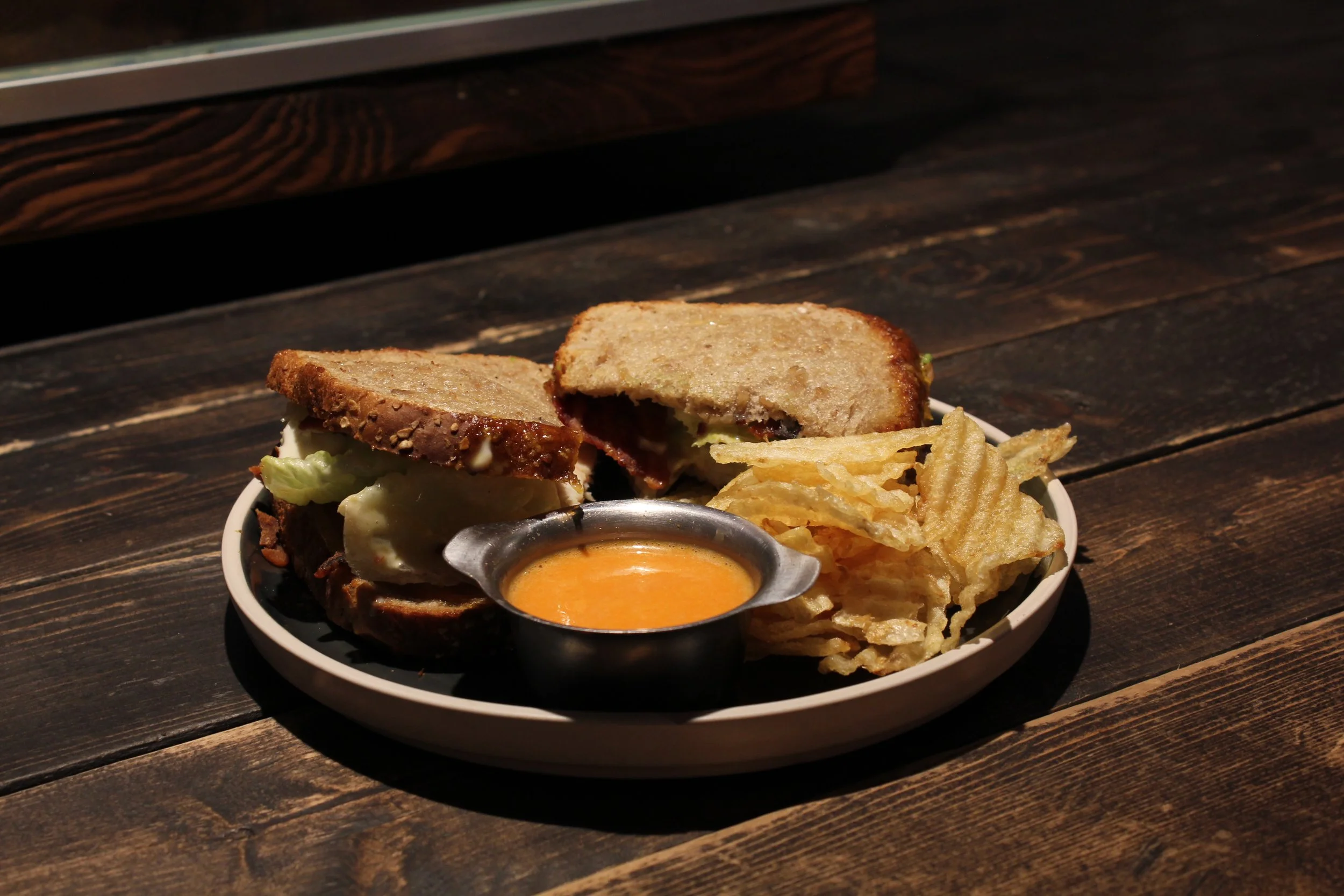A plate with a sandwich, potato chips, and a small container of orange dipping sauce on a wooden table.