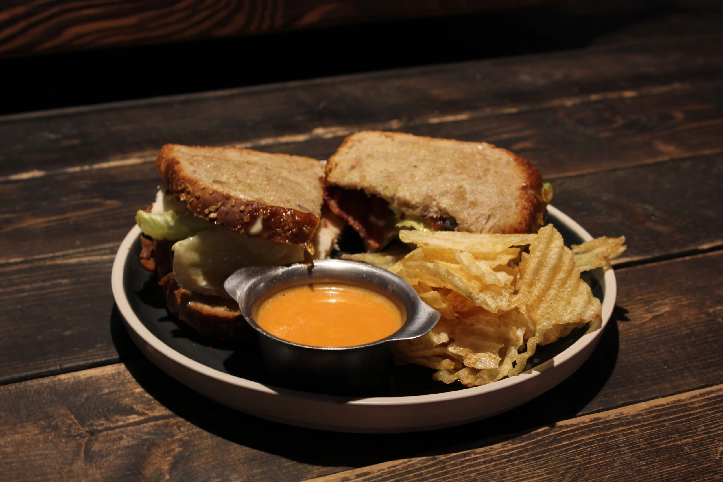 A plate with a sandwich, potato chips, and dipping sauce on a wooden table.