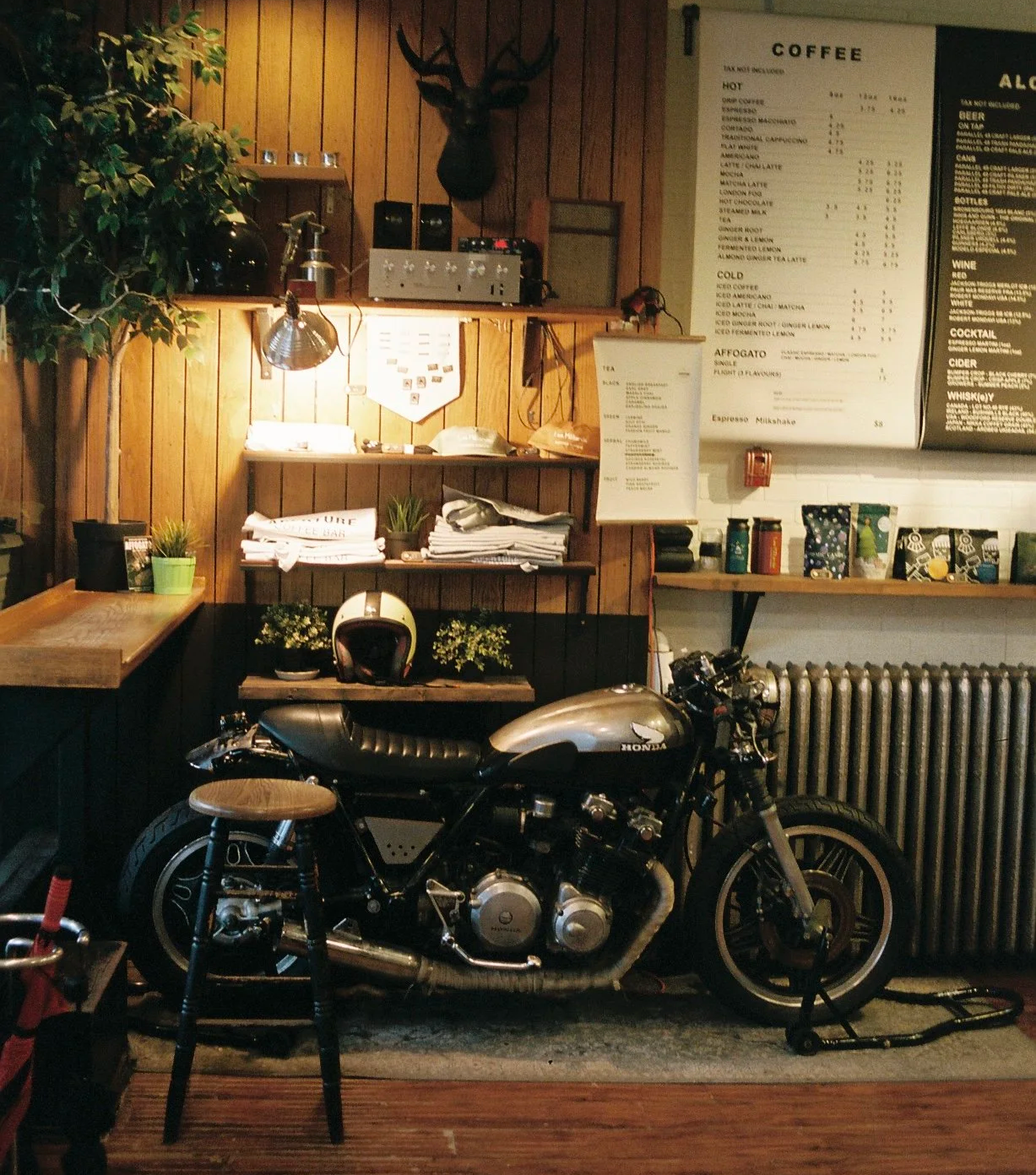 A silver vintage Honda motorcycle parked indoors in a coffee shop with wooden wall paneling, potted plants, stacked newspapers, a helmet, a small stool, and a menu board.