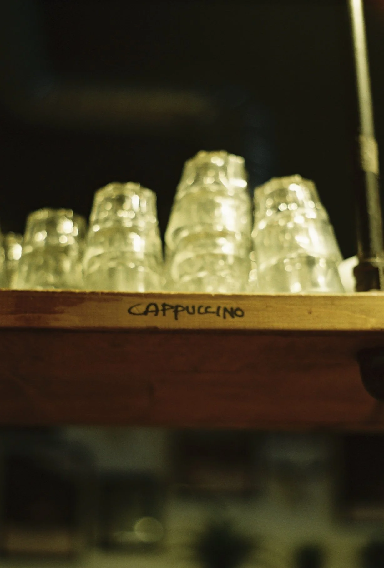 Stacked glass cups labeled 'Cappuccino' on a wooden shelf in a dimly lit setting.