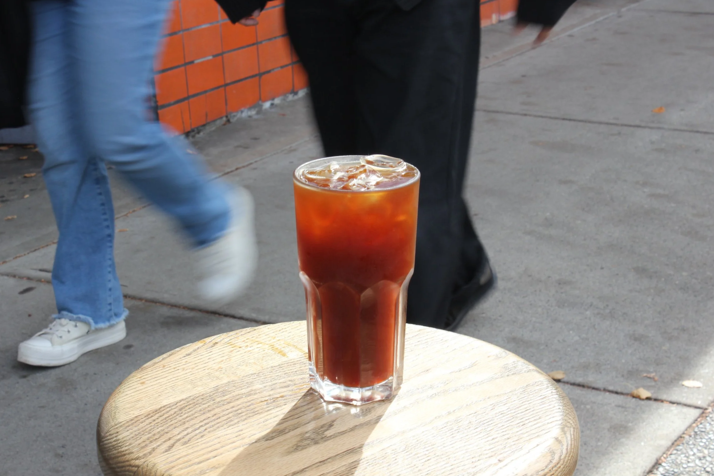 A tall glass of iced coffee on a small wooden table outside on a sidewalk, with people walking by in the background.