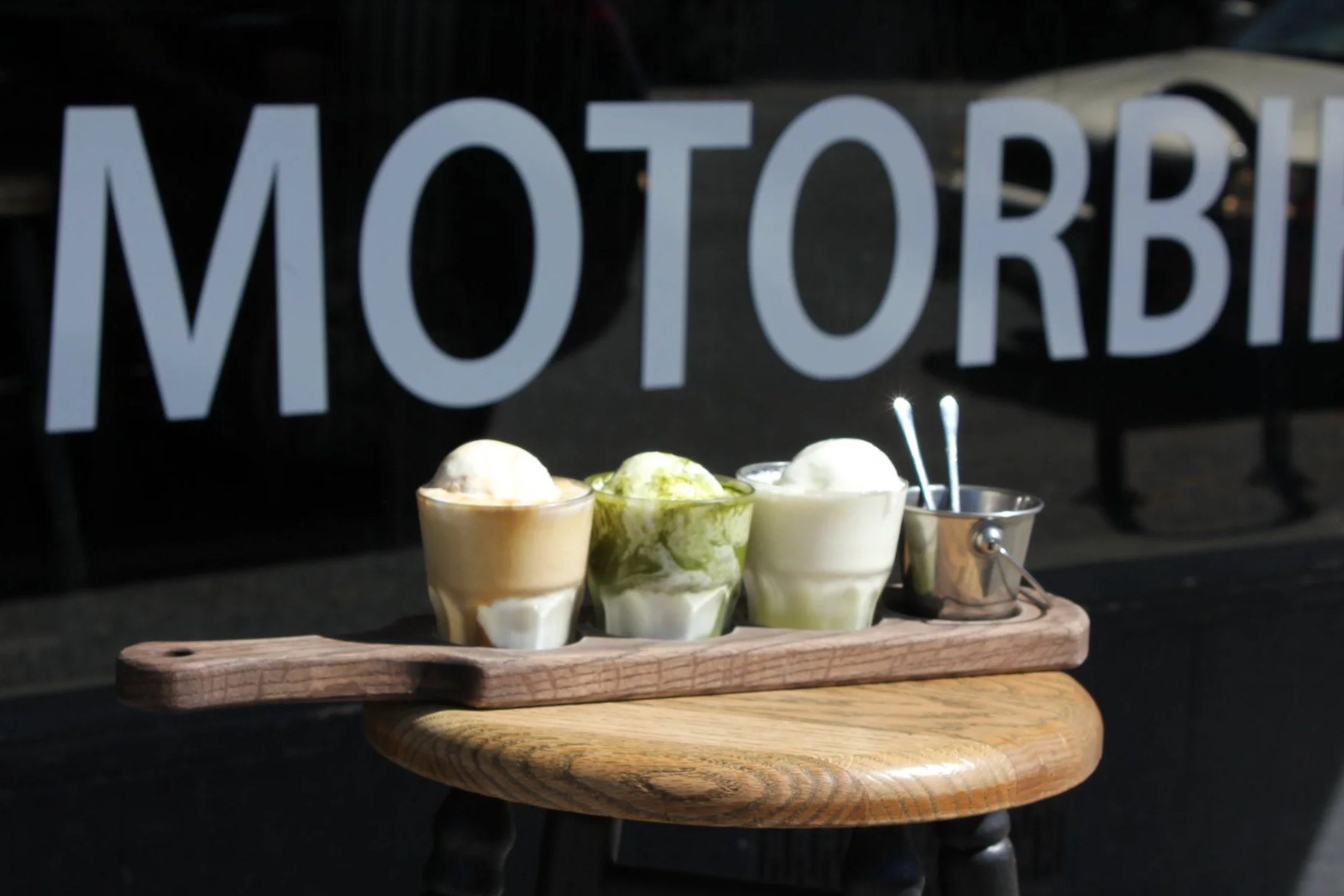 Three glasses of ice cream in front of a window with 'MOTORBI' written on it, placed on a wooden table with a small metal bucket and two silver spoons.