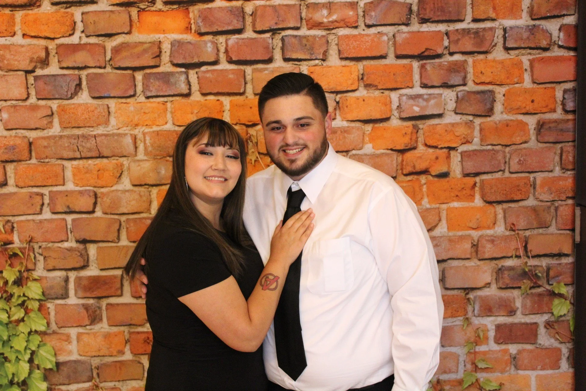 A woman and man smiling and posing together in front of a brick wall. The woman has long brown hair, wearing a black shirt, and has a tattoo on her left arm. The man has short dark hair, a beard, and is dressed in a white shirt with a black tie.