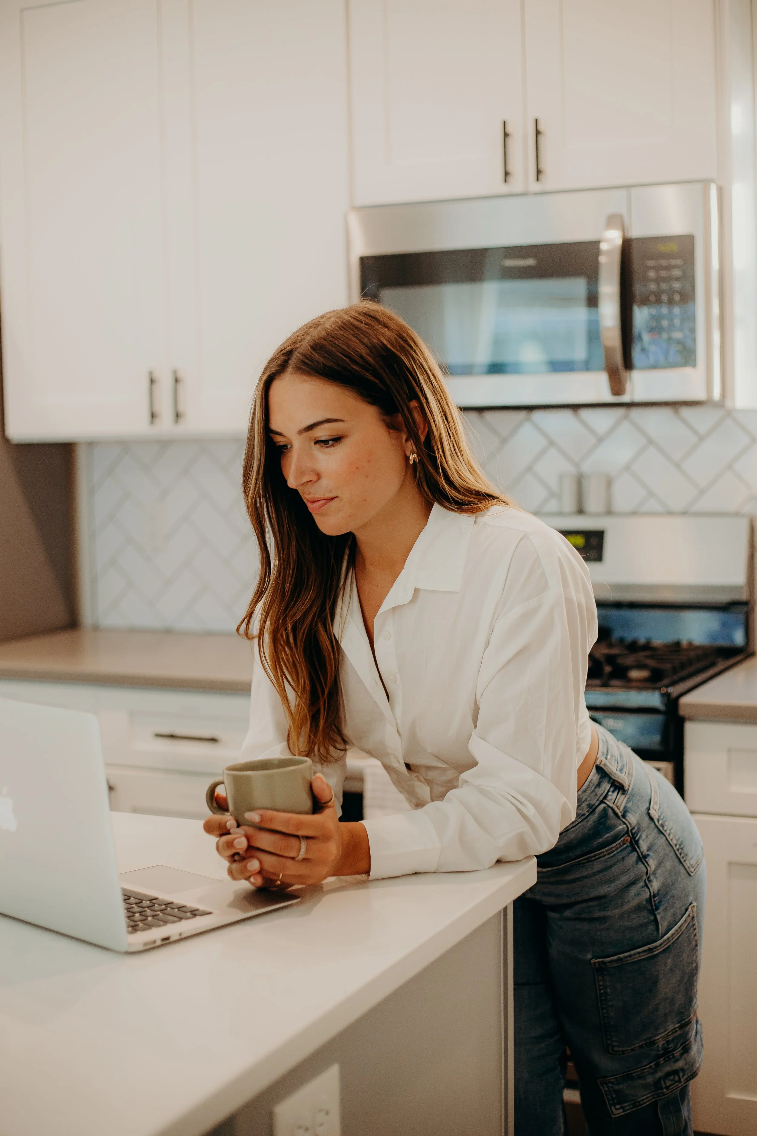 A young woman with long brown hair, wearing a white shirt and jeans, leaning on a kitchen counter holding a mug, looking at an open laptop.