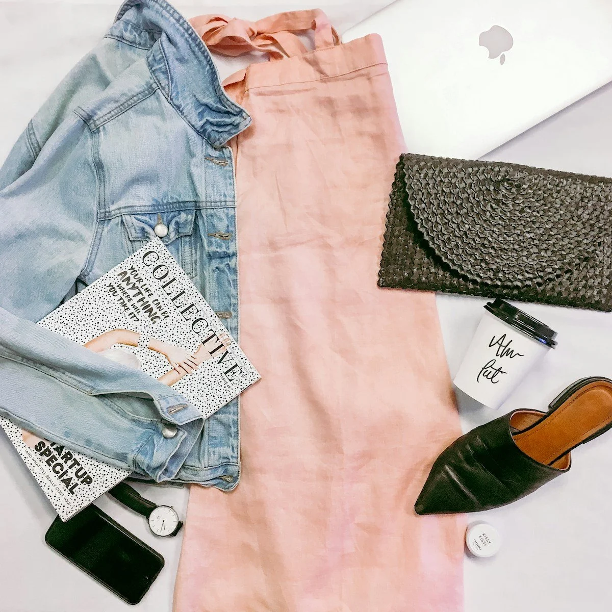 Flat lay of fashion and workspace items including a denim jacket, pink skirt, black textured clutch, coffee cup with 'Will & Grace' written, black pointed shoe, white Lip Balm, smartphone, wristwatch, and a laptop in the background.