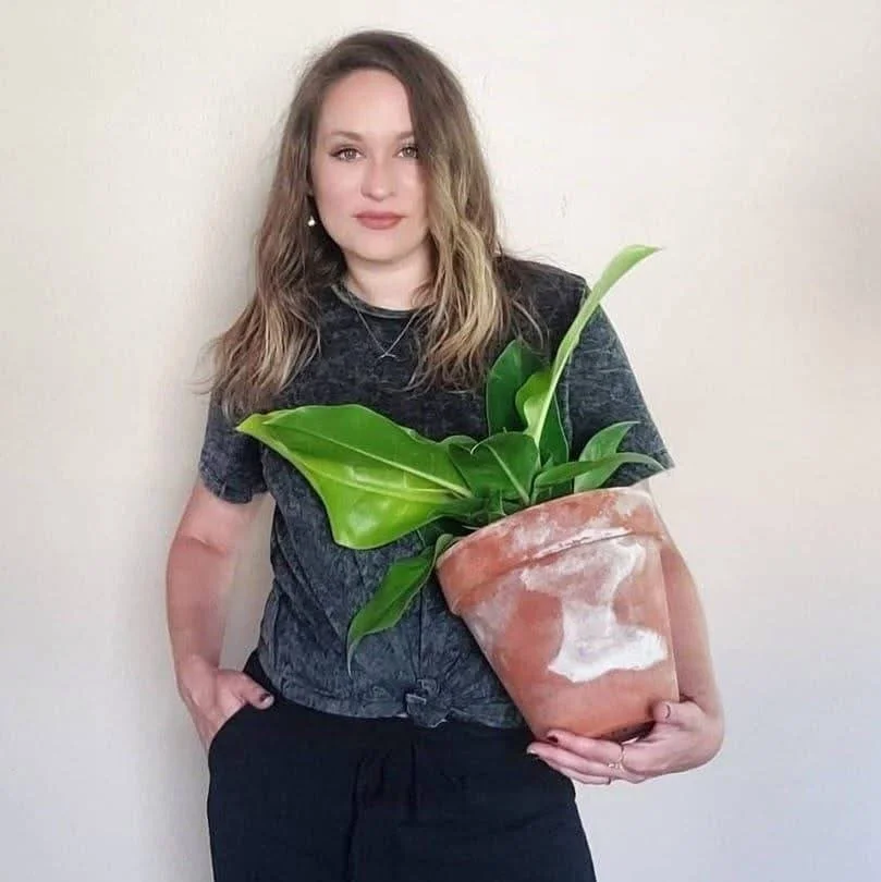 A woman with wavy brown hair wearing a dark gray t-shirt and black pants, holding a large potted plant with broad green leaves, standing against a plain off-white wall.