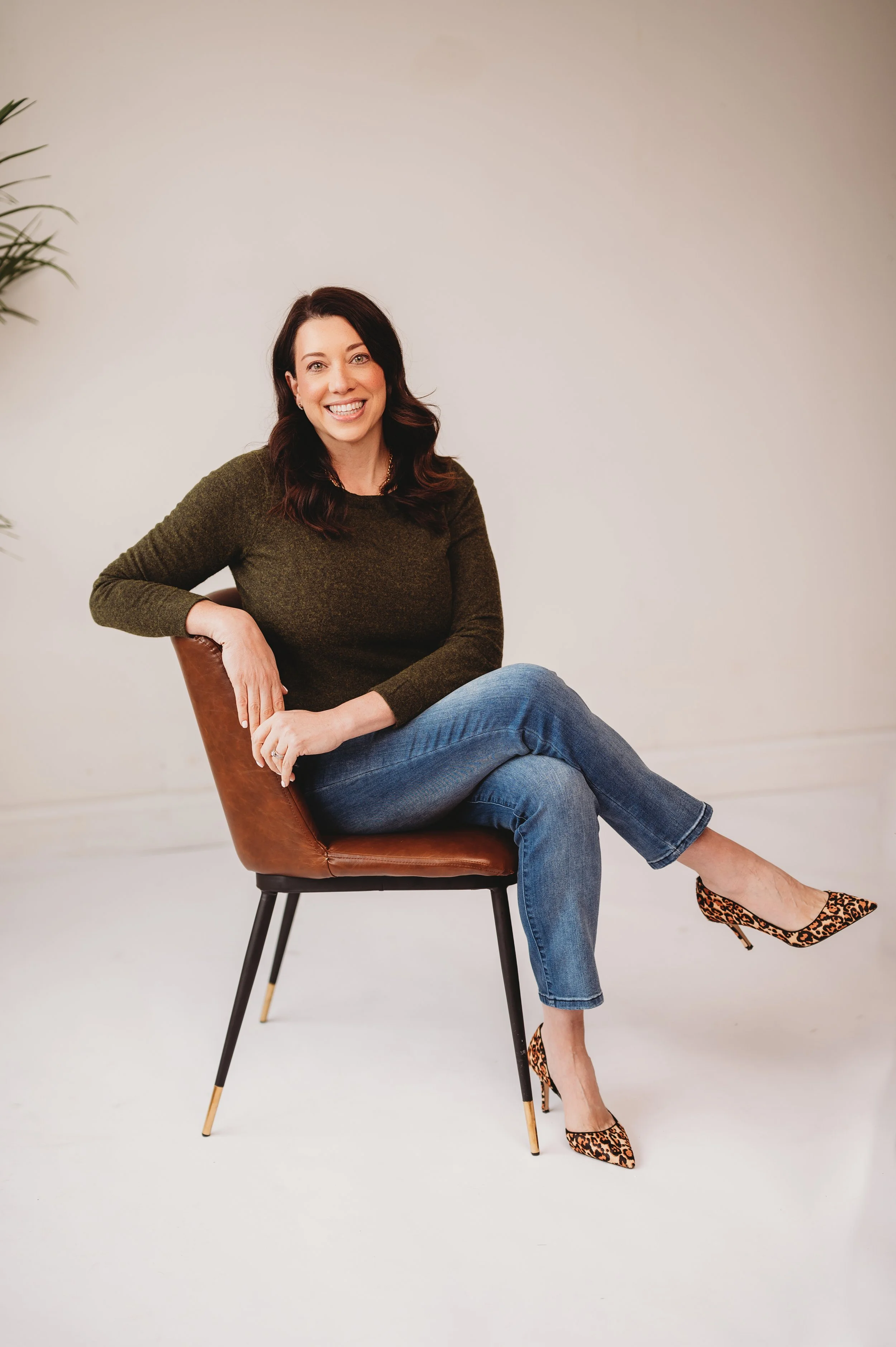 Natalie Ranalli sitting on a brown chair against a plain light-colored background, smiling at the camera.