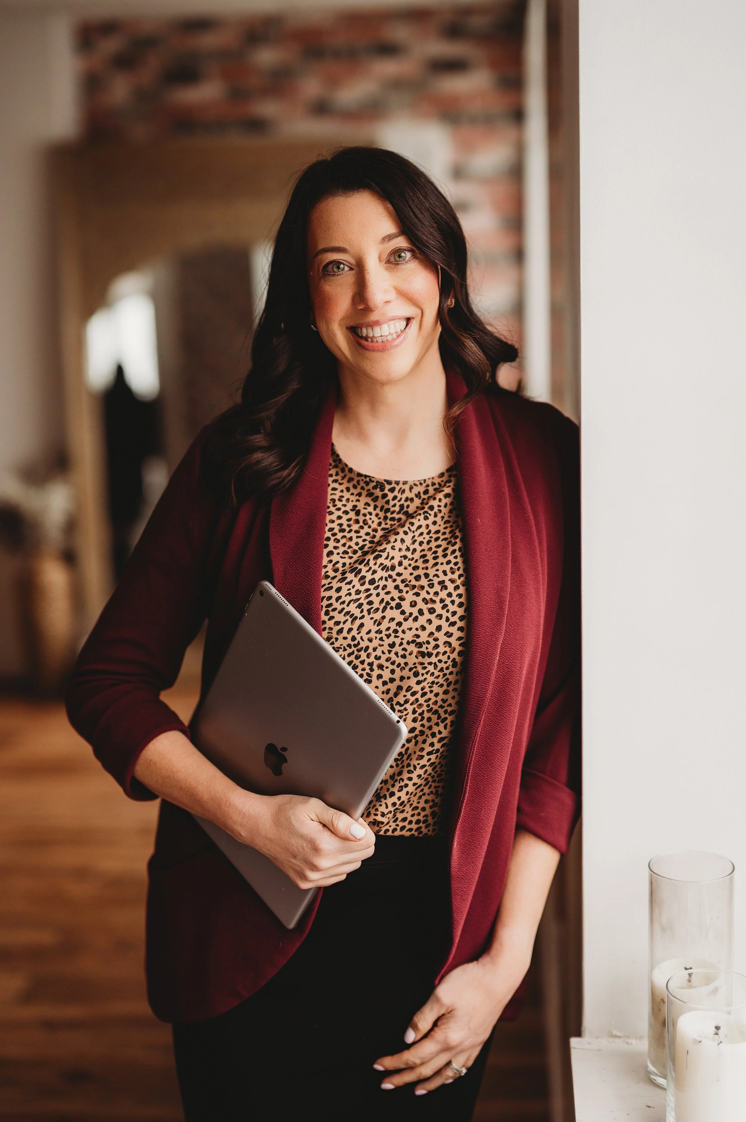 Natalie Ranalli smiling and holding a closed silver MacBook, standing in a cozy indoor setting with a brick wall background and candles on a windowsill.