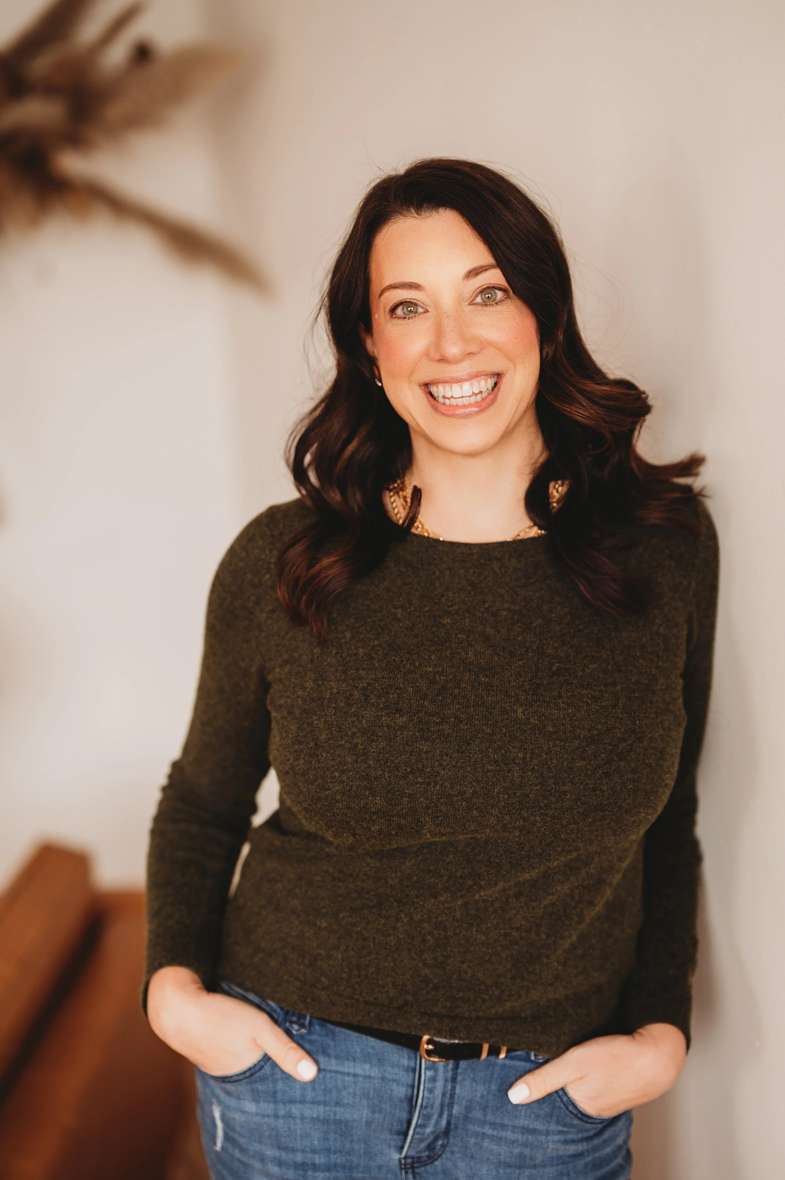 Natalie Ranalli standing against a plain wall, wearing a dark green sweater, blue jeans, and a gold necklace.