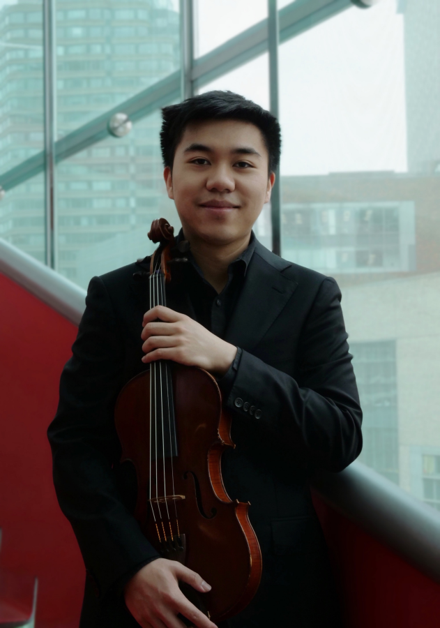 Young man in a black suit holding a violin on a balcony with city buildings and large windows in the background.