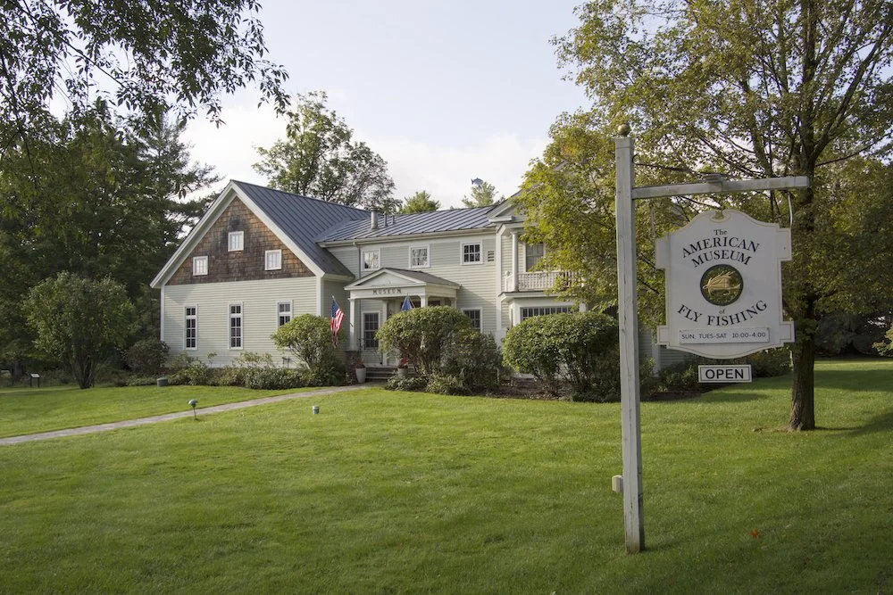 A large white house with a grey roof, surrounded by green trees and shrubs, has a sign in the front yard that reads 'The American Museum of Fly Fishing.' The sign indicates the museum is open and open hours are Sunday through Saturday from 10 am to 4 pm.