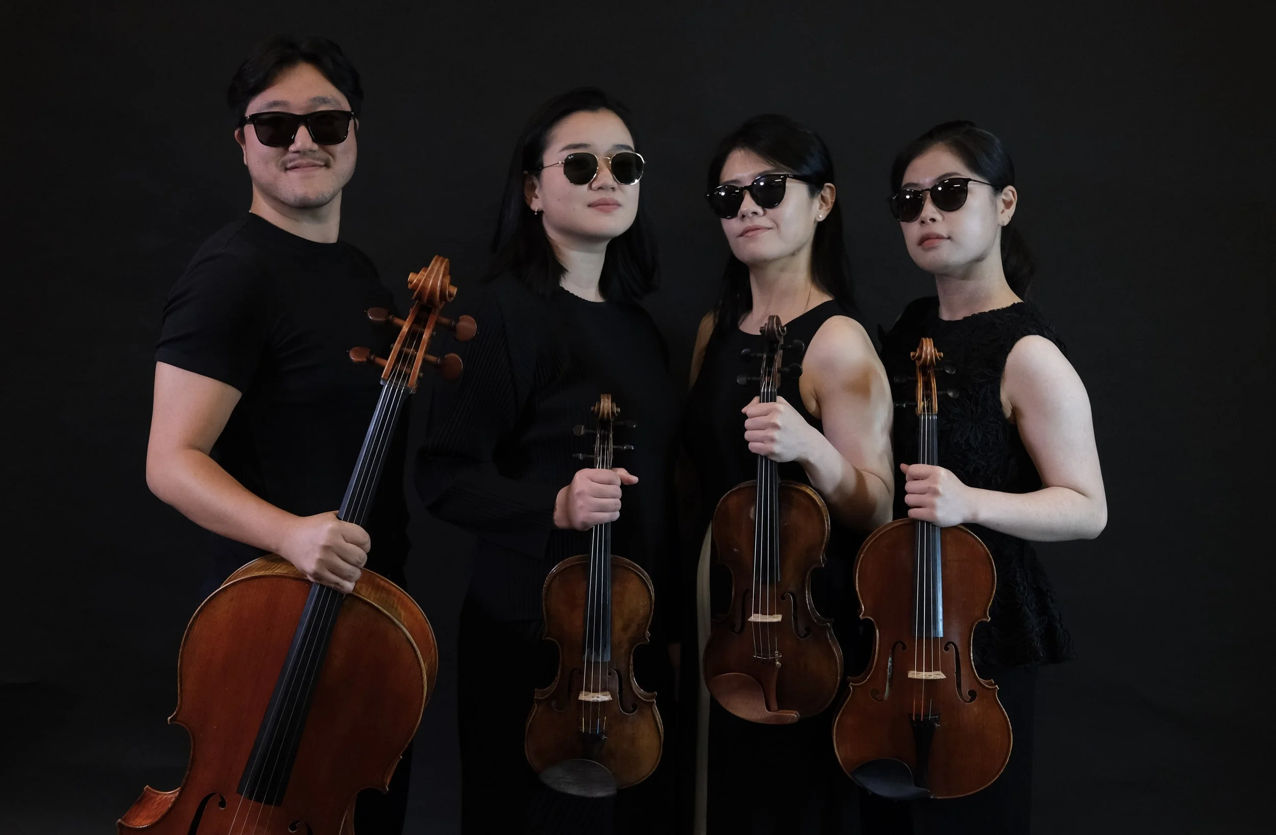 Four musicians holding violins and a cello, wearing black clothing and sunglasses, standing in front of a black background.