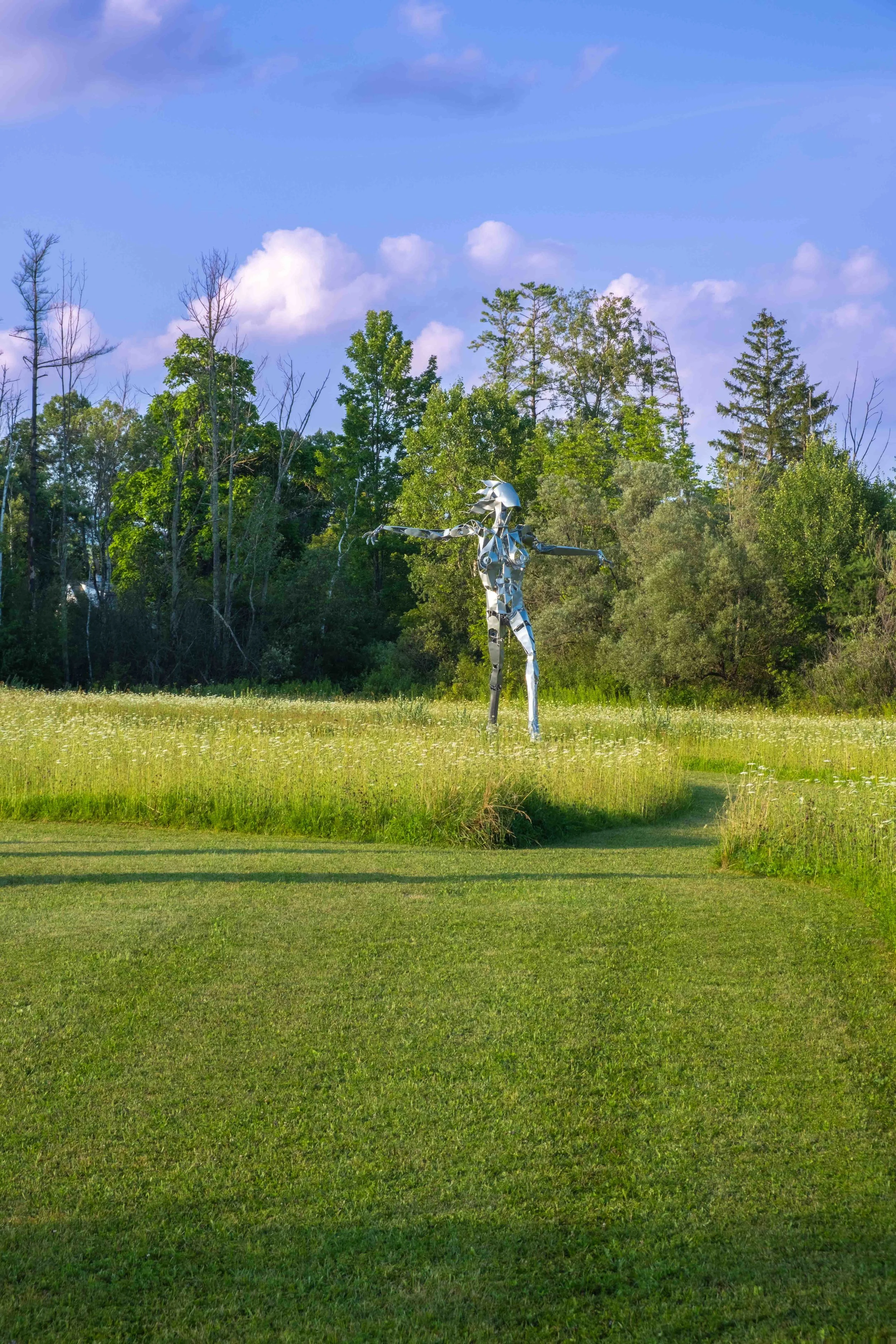 A metallic humanoid sculpture with outstretched arms standing in a grassy field with trees and blue sky in the background.