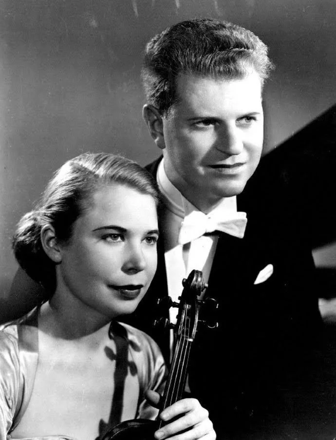 A black and white photo of a young man and woman dressed formally, the woman holding a violin. The woman has short styled hair, and the man has a pompadour hairstyle and is wearing a tuxedo with a bow tie.