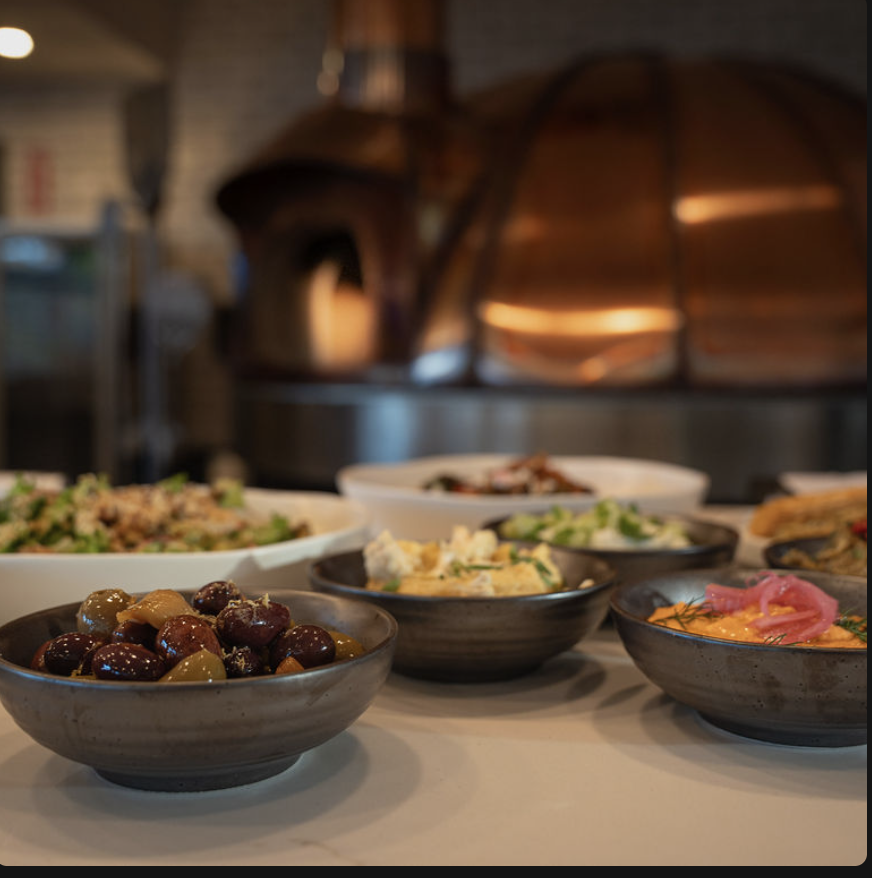 Various dishes of food on a restaurant table, including salads, olives, and dips, with a wood-fired oven in the background.