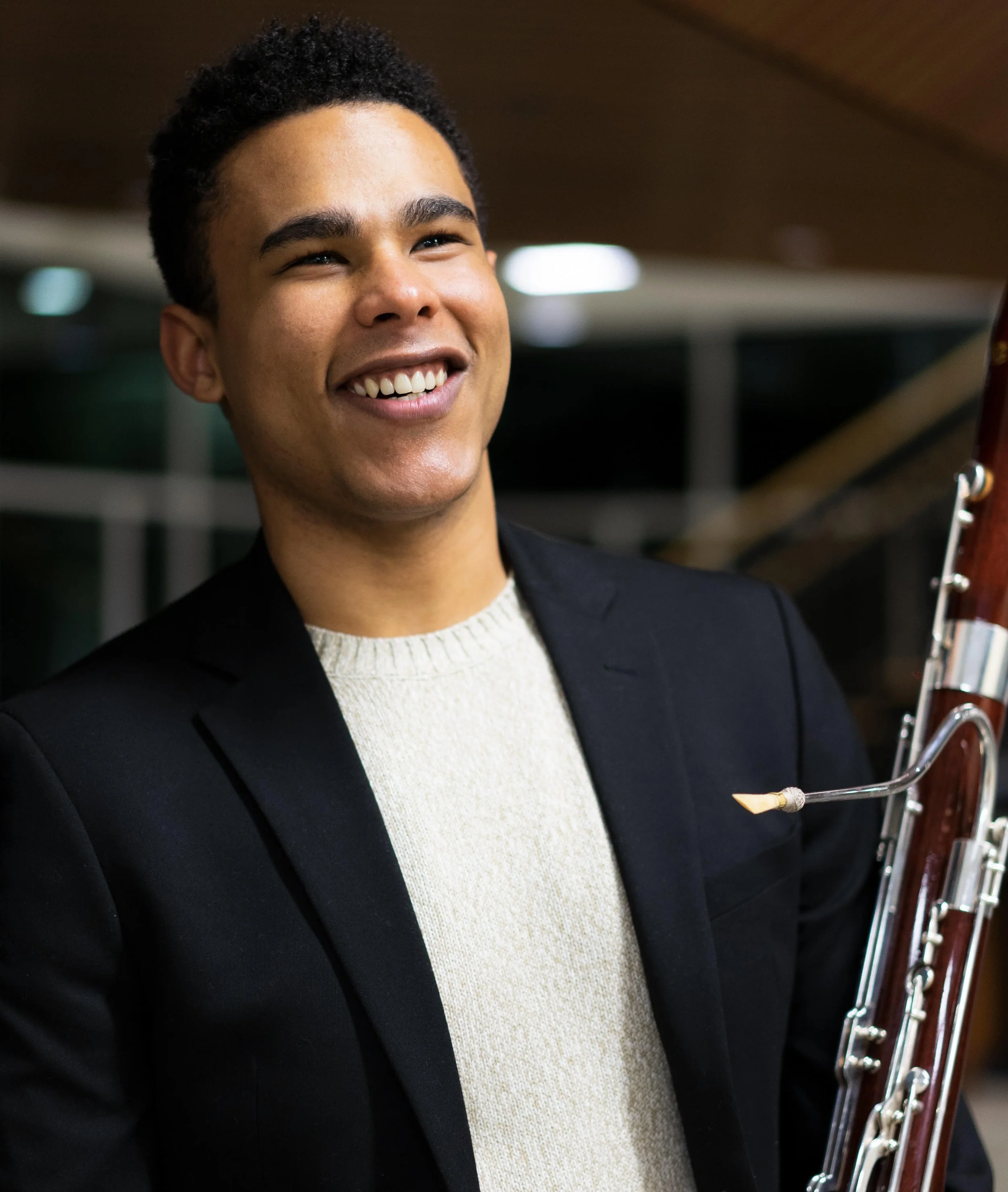 A young man smiling, holding a bassoon, dressed in a black blazer and cream sweater, in an indoor setting.
