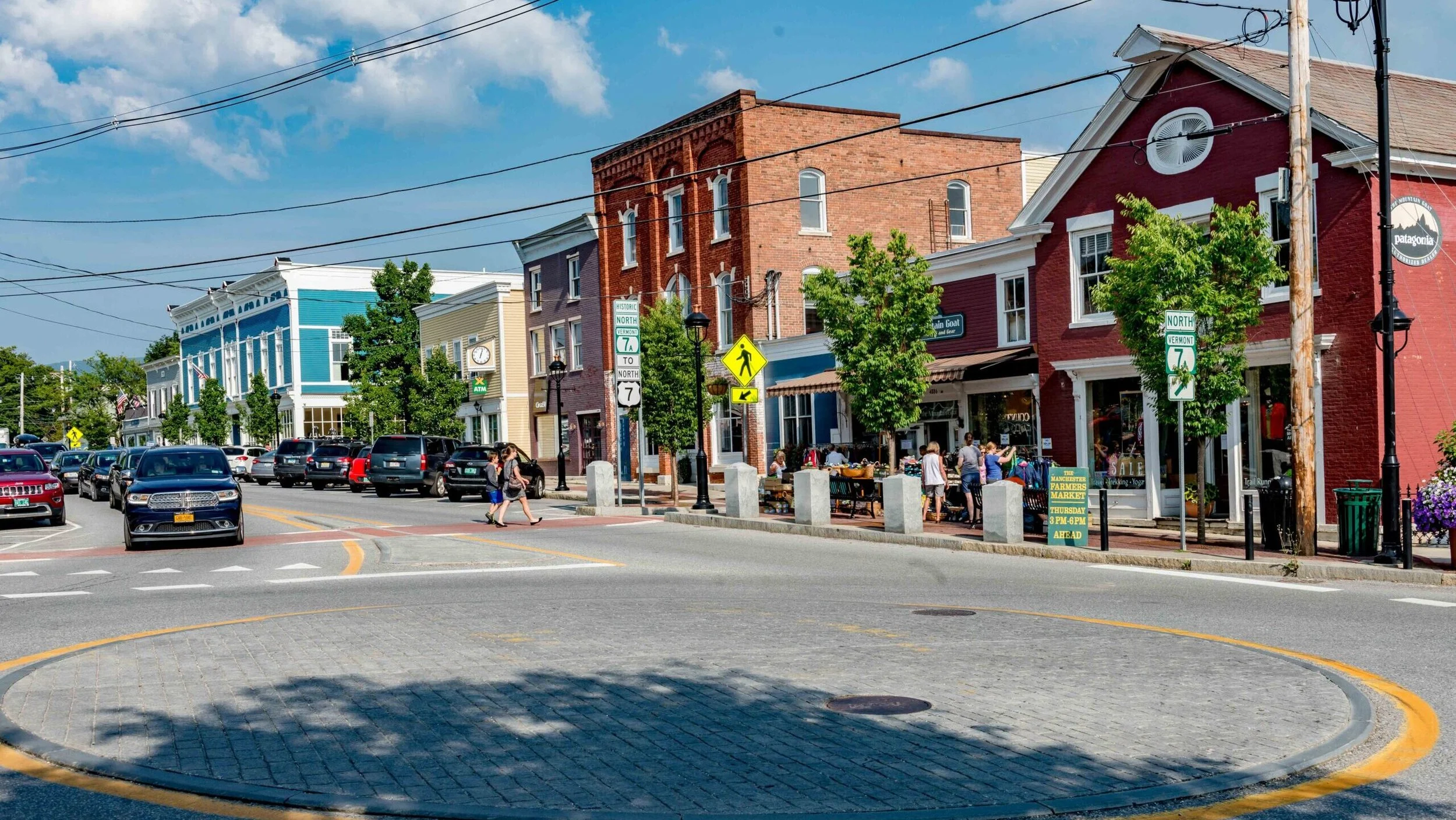 A small town street with colorful buildings, parked cars, pedestrians, and trees. The buildings are painted in shades of blue, yellow, and red, with storefronts and signs indicating shops and markets.