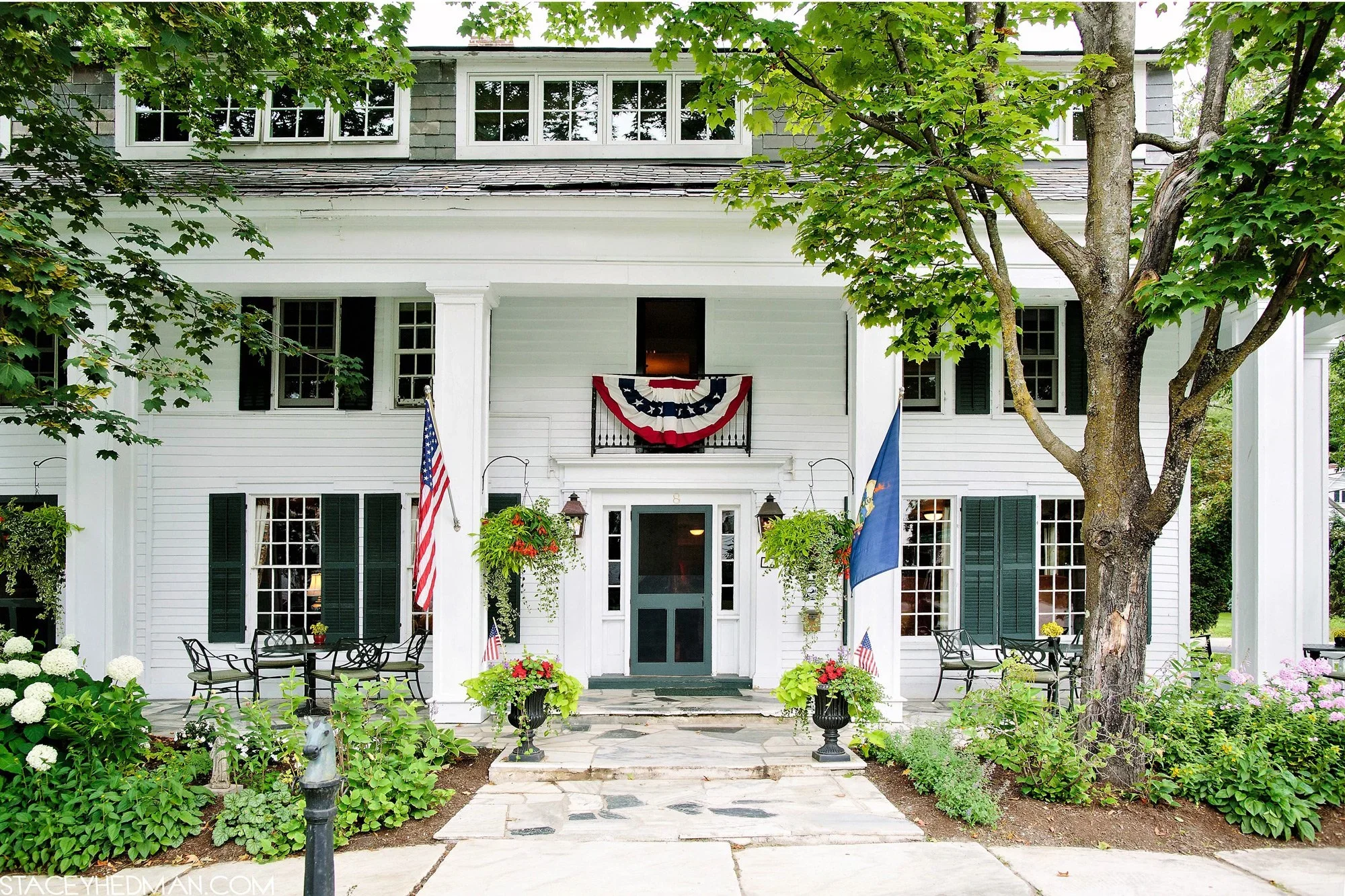 Front view of a white house decorated with American flags, a patriotic bunting, and hanging flower baskets, with a tree and garden in the foreground.