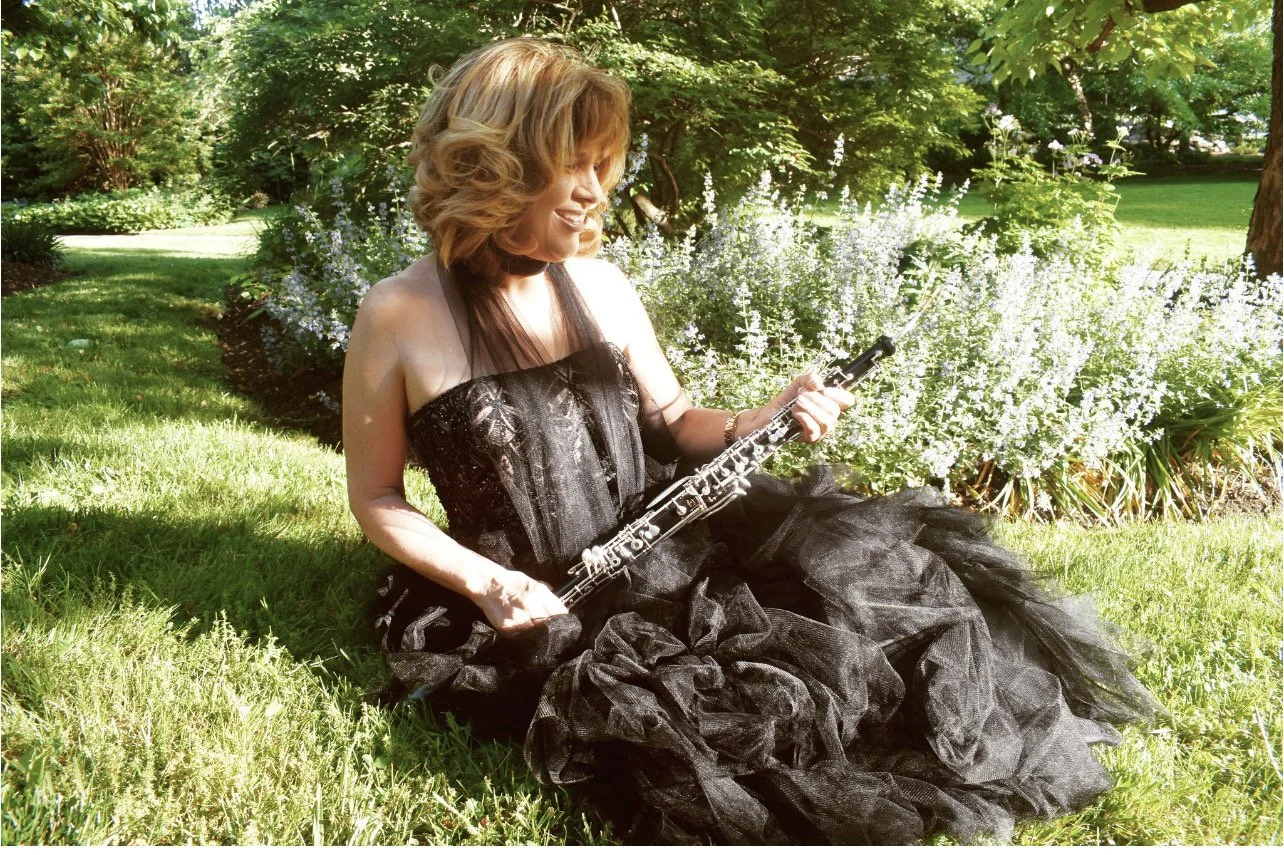 A woman in a black lace dress sitting on grass in a garden, holding a clarinet, smiling, surrounded by greenery and white flowers.