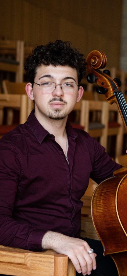 Young man with curly hair and glasses holding a cello in a room with wooden chairs.