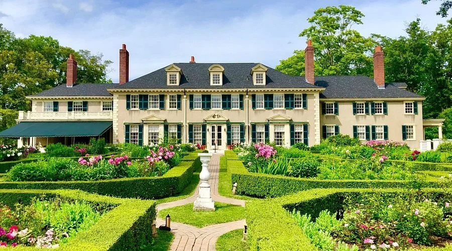 Large historic mansion with multiple chimneys and dormer windows, surrounded by a manicured garden with greenery and pink flowers.