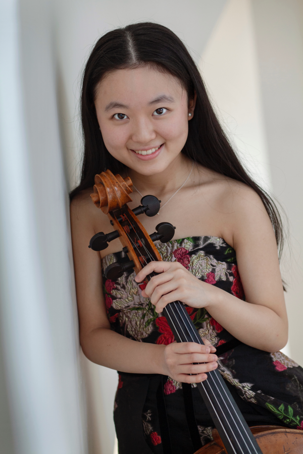 A young woman with long dark hair smiling and holding a violin, wearing a strapless dress with floral patterns.