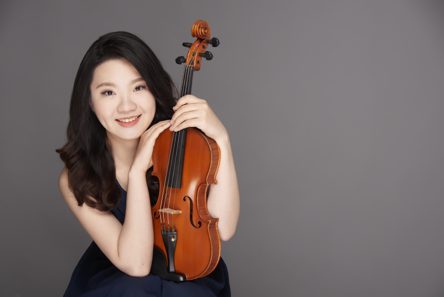 A young woman with long, dark hair smiles while holding a violin against a gray background.