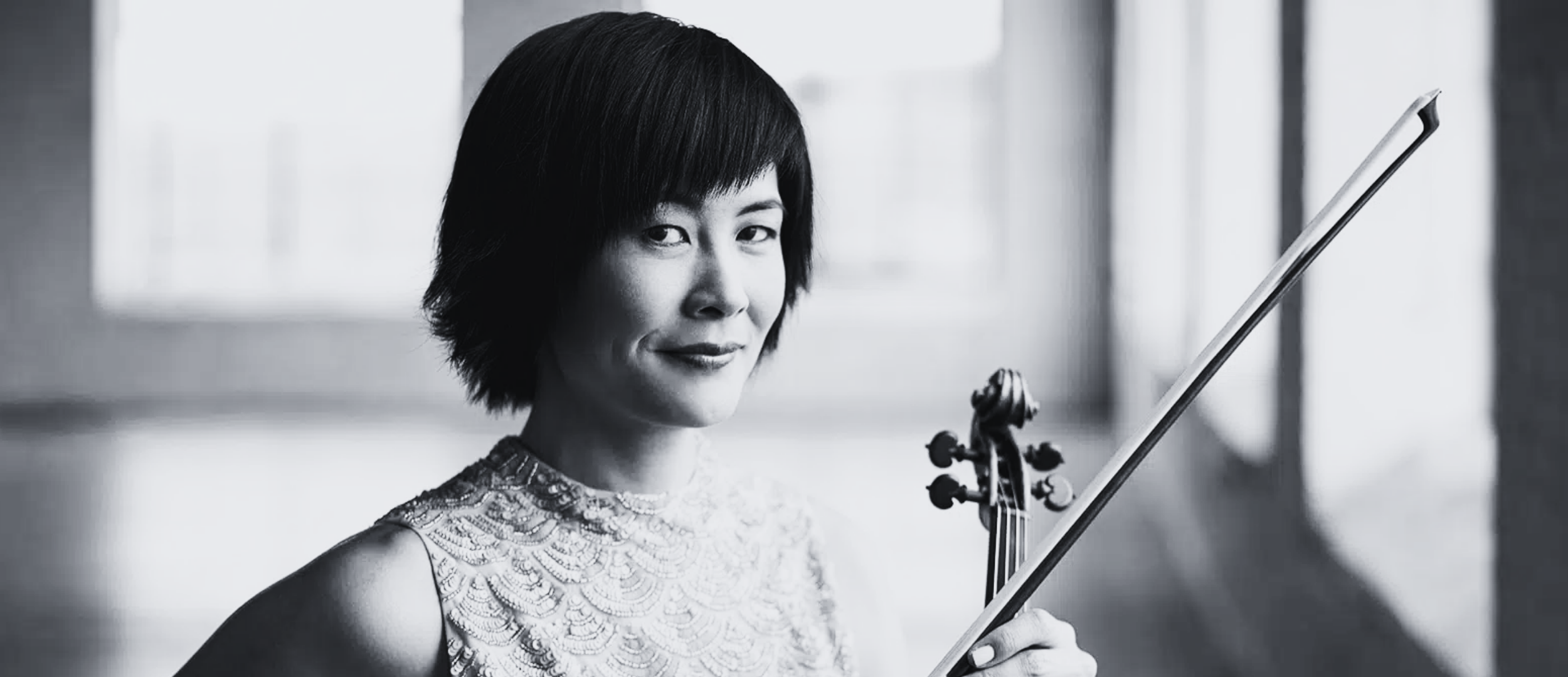 Black and white photo of a woman holding a violin, smiling, with a lace top, standing indoors near a window.