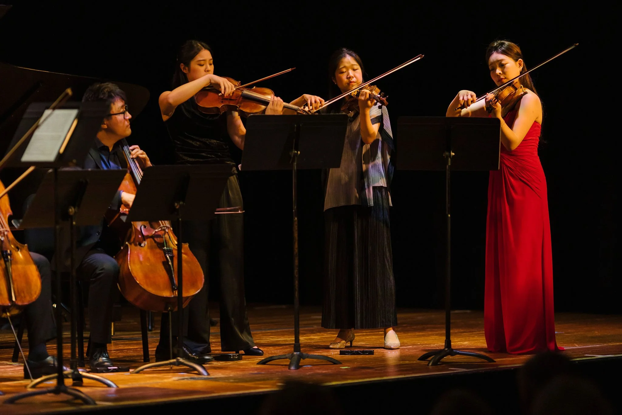 Orchestra musicians performing on stage, including three women playing violins, one woman playing a cello, and a man playing the piano.