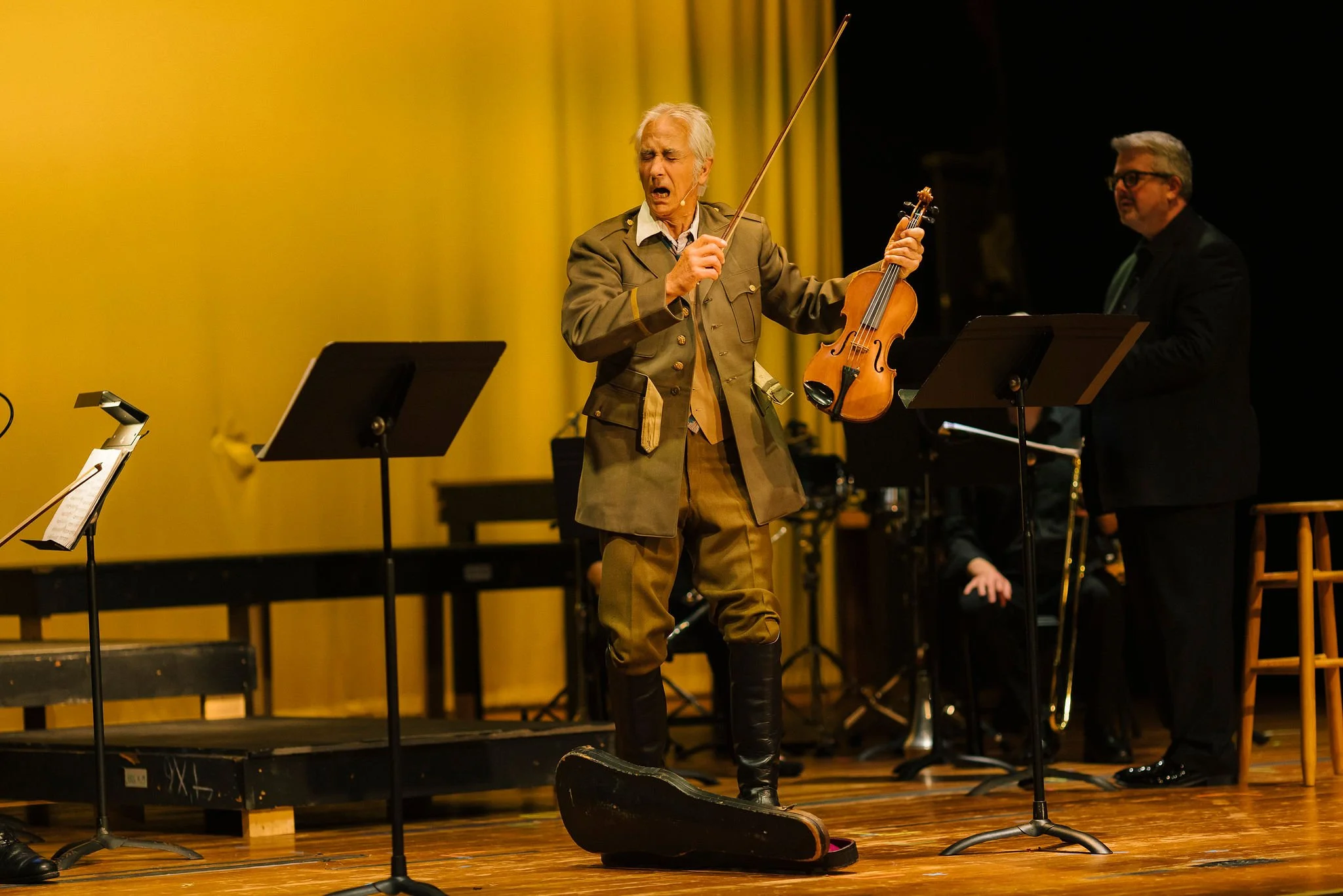 A man in historic costume performing with a violin on stage, with a music stand and band members in the background.